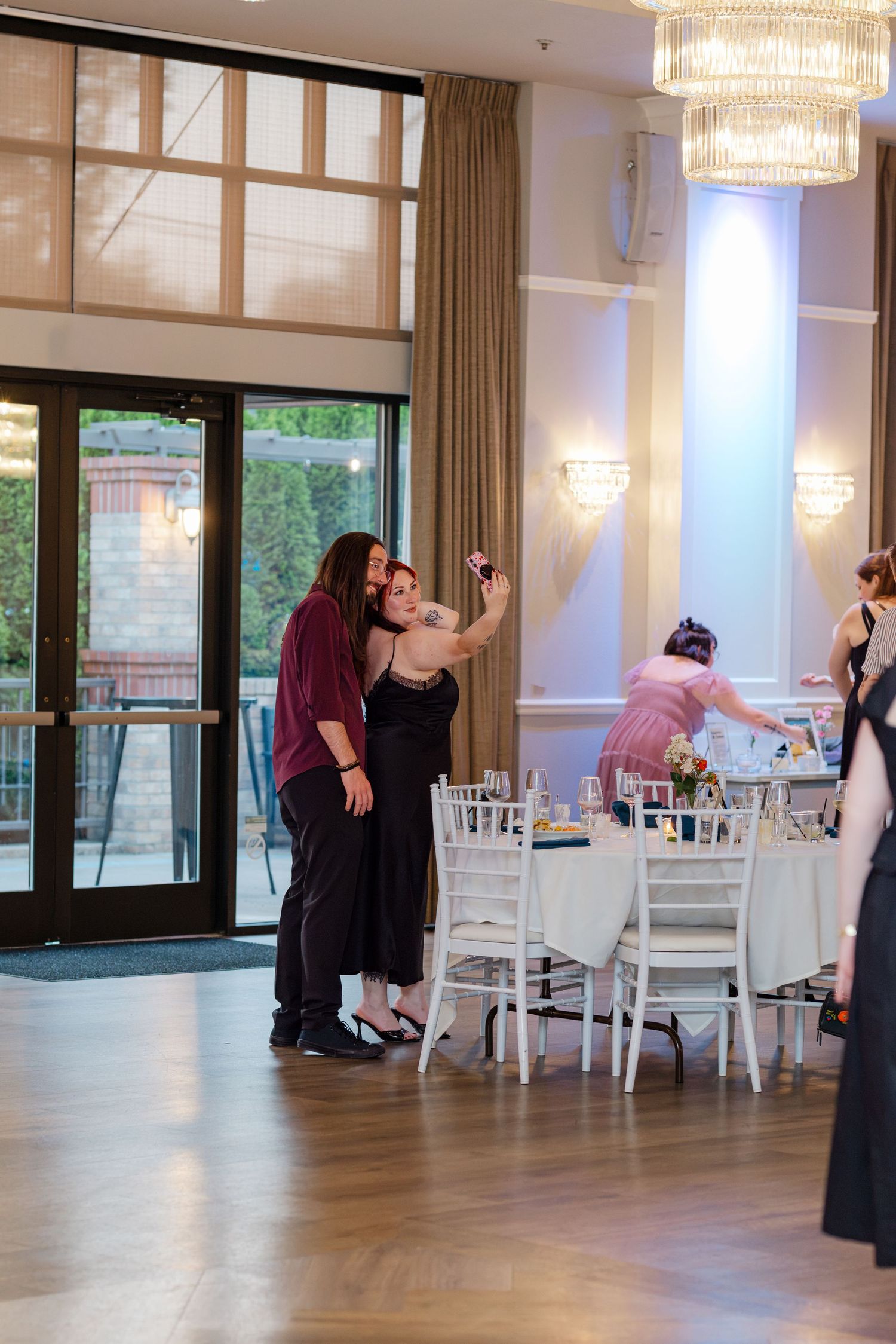 Two wedding guests take a selfie on the dance floor of an elegant chandelier-lit reception venue.