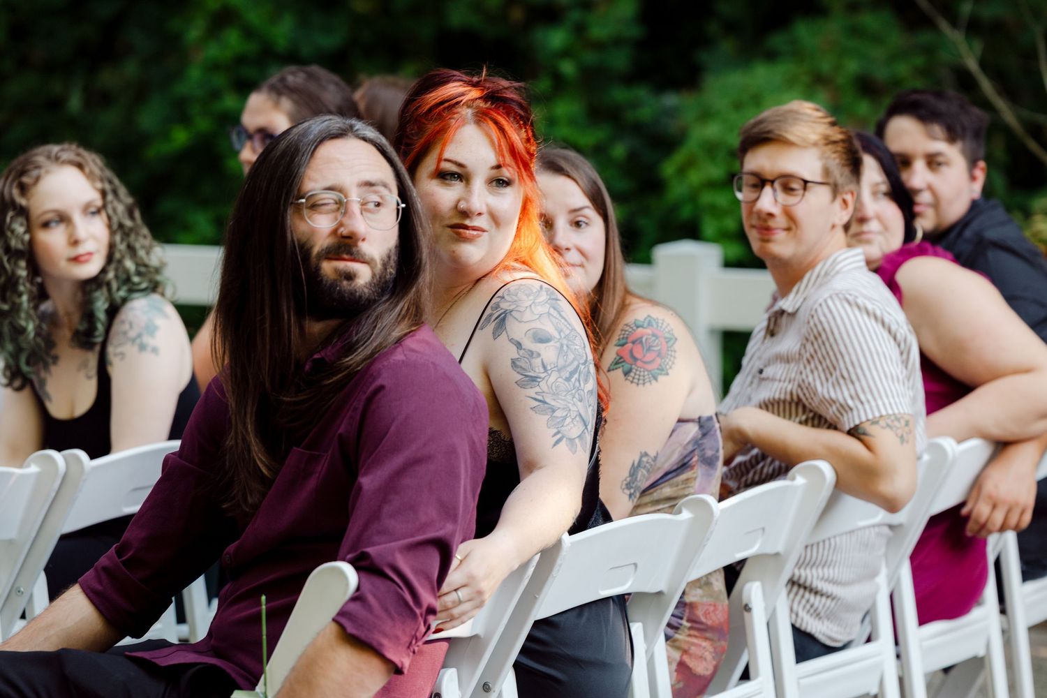Wedding guests with colorful hair and tattoos seated in white chairs at an outdoor ceremony.