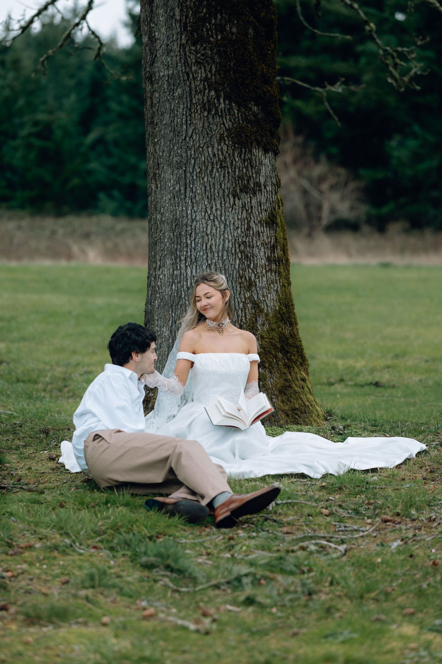 A couple relaxes under a large tree in a pastoral setting during a romantic photoshoot.