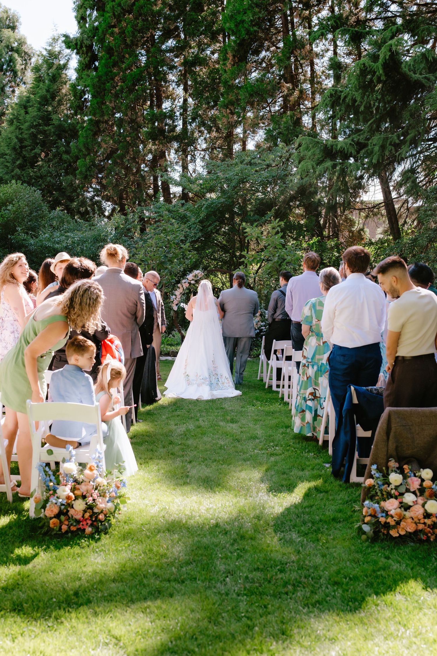Outdoor wedding ceremony on green lawn with guests standing along flower-lined aisle as bride walks toward floral arch.