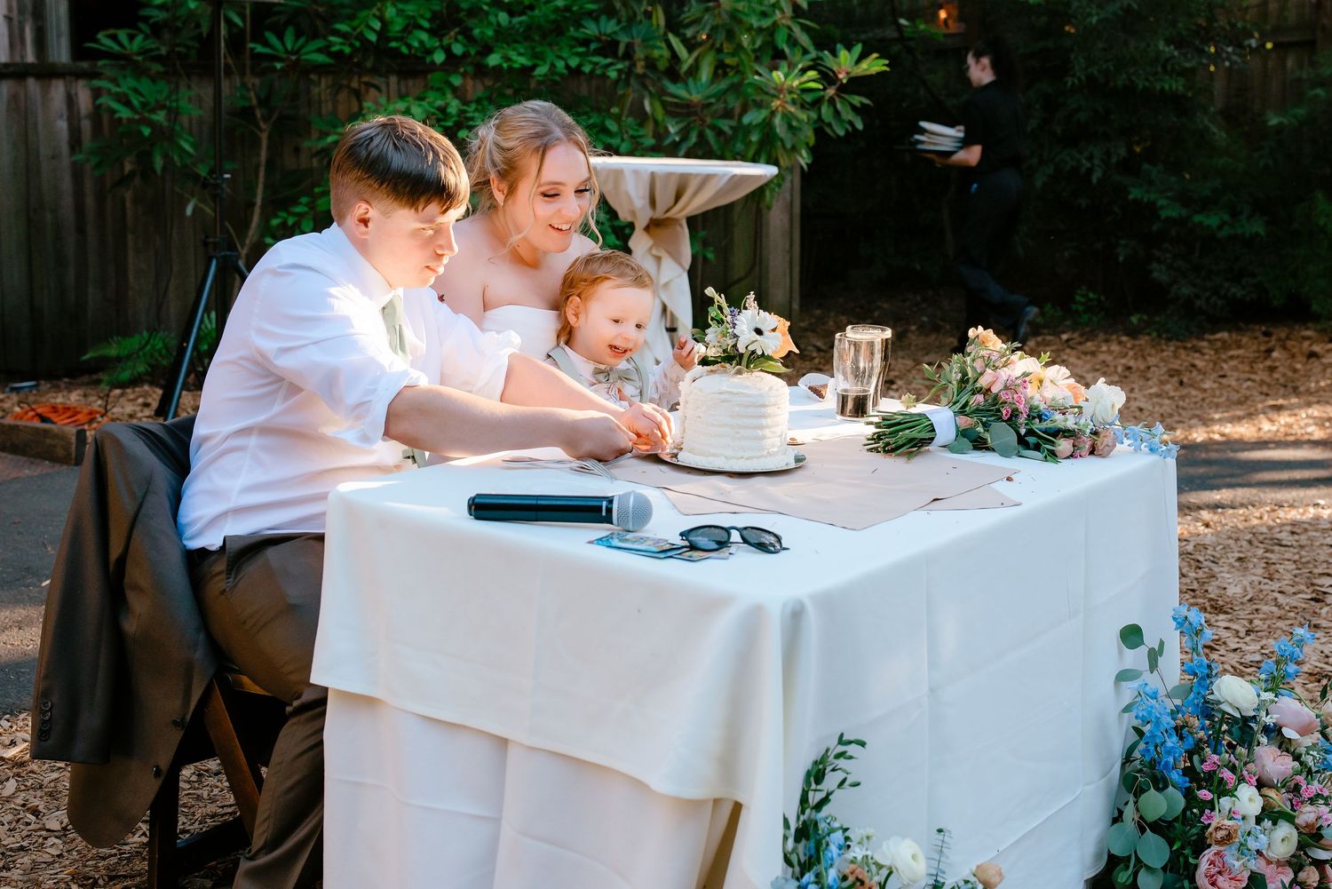 Bride in white gown cuts wedding cake with young child and teen at reception table.