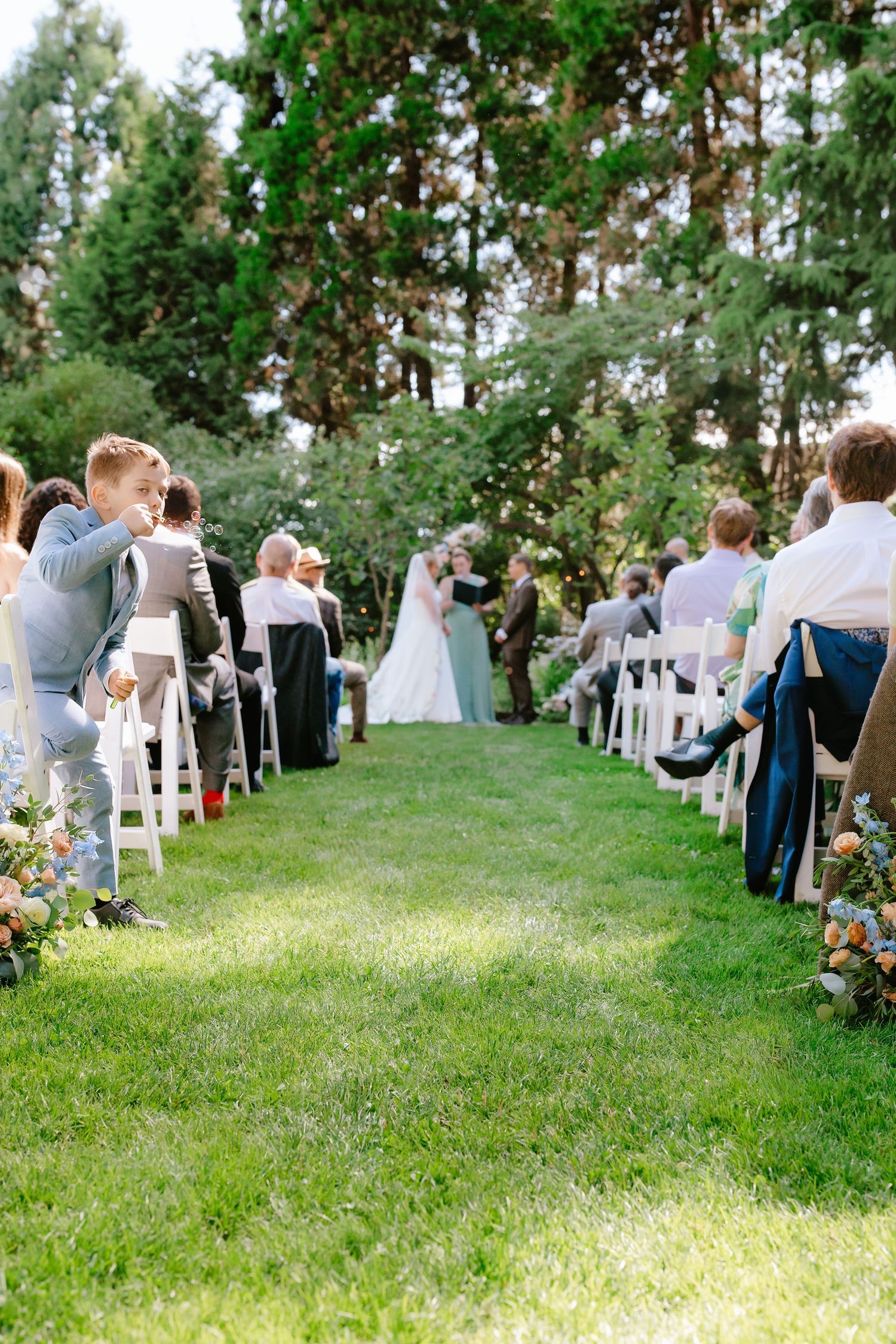Outdoor wedding ceremony on green lawn with guests seated in white chairs, bride and groom at altar surrounded by tall trees.