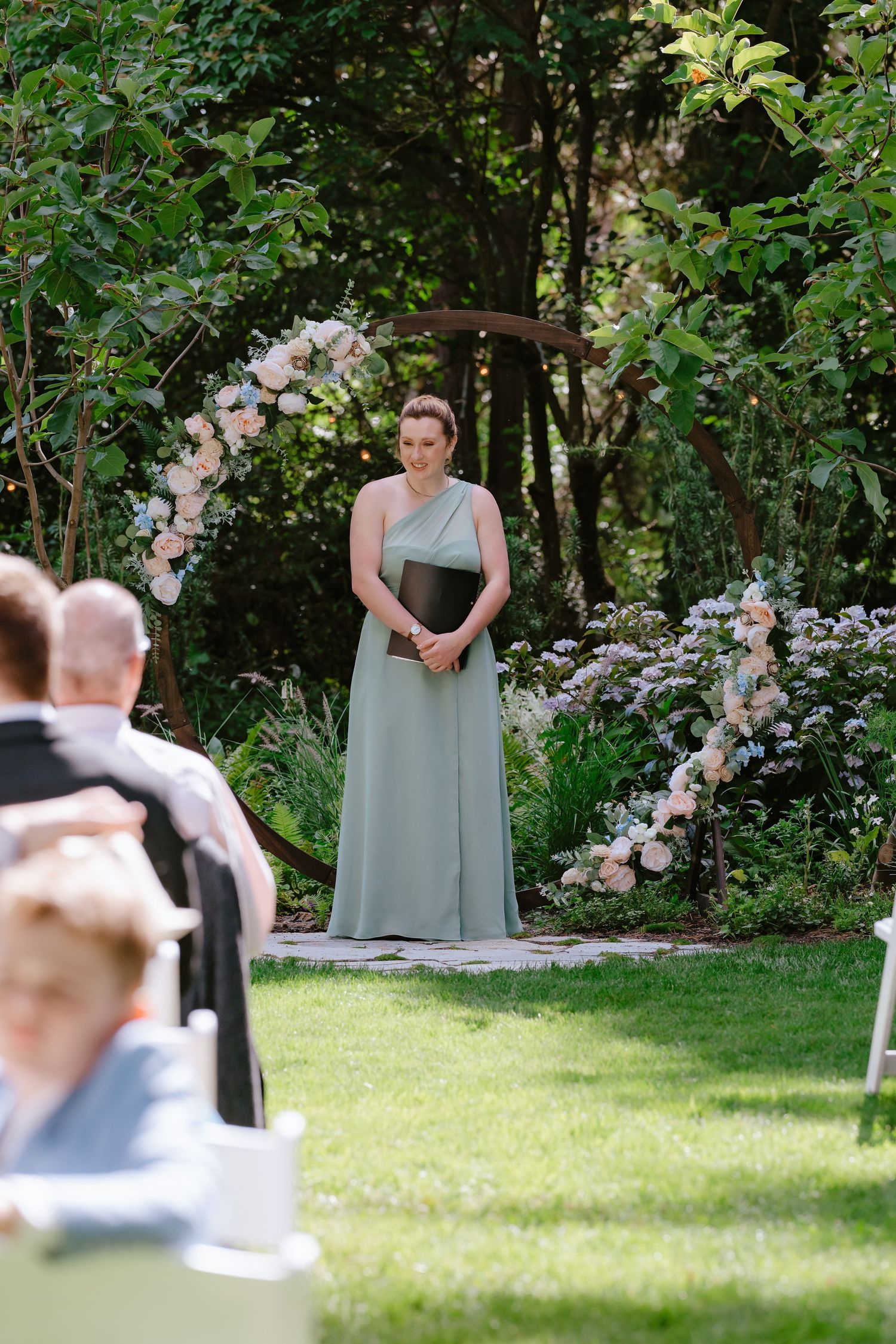A woman in a sage green bridesmaid dress stands at a floral wedding arch in a garden ceremony.