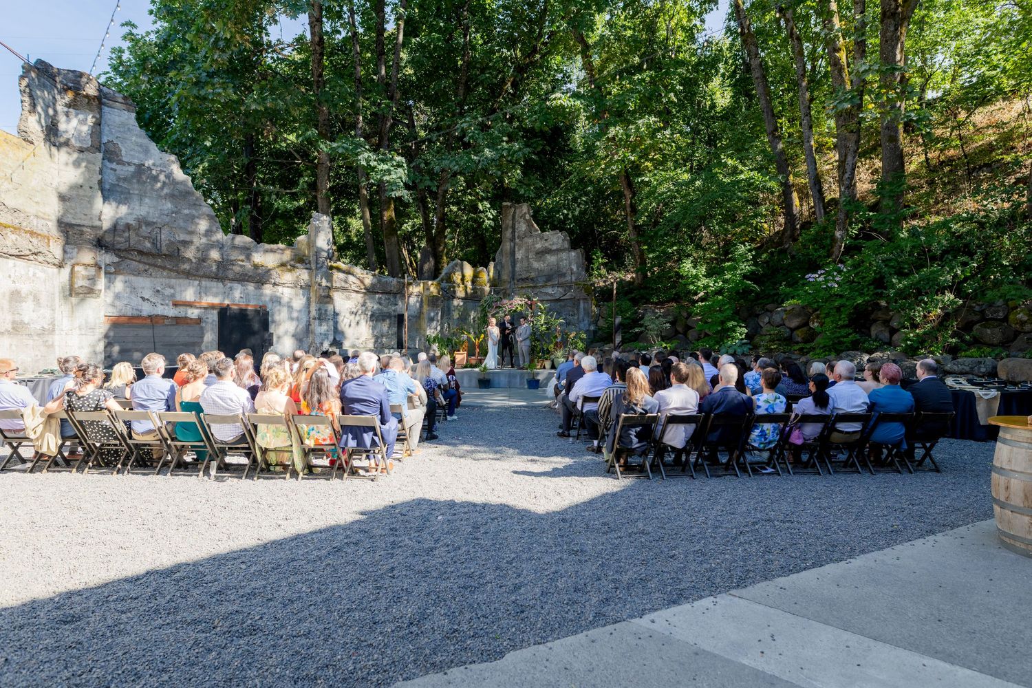 Outdoor wedding ceremony with guests seated in rows amid ancient stone ruins surrounded by lush green trees.