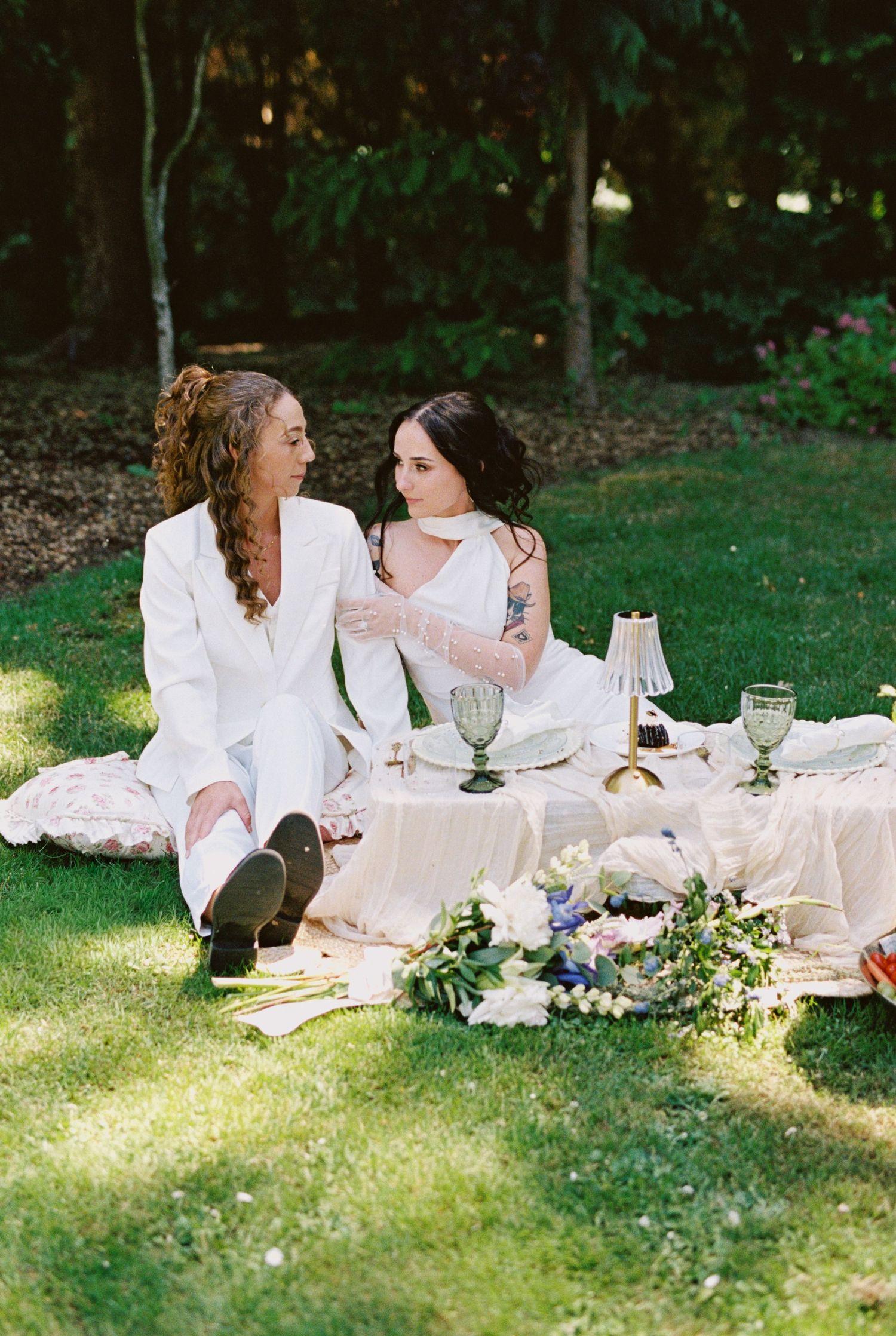 Two brides in white sit on grass at an elegant outdoor picnic wedding setup with floral arrangements and vintage glassware.