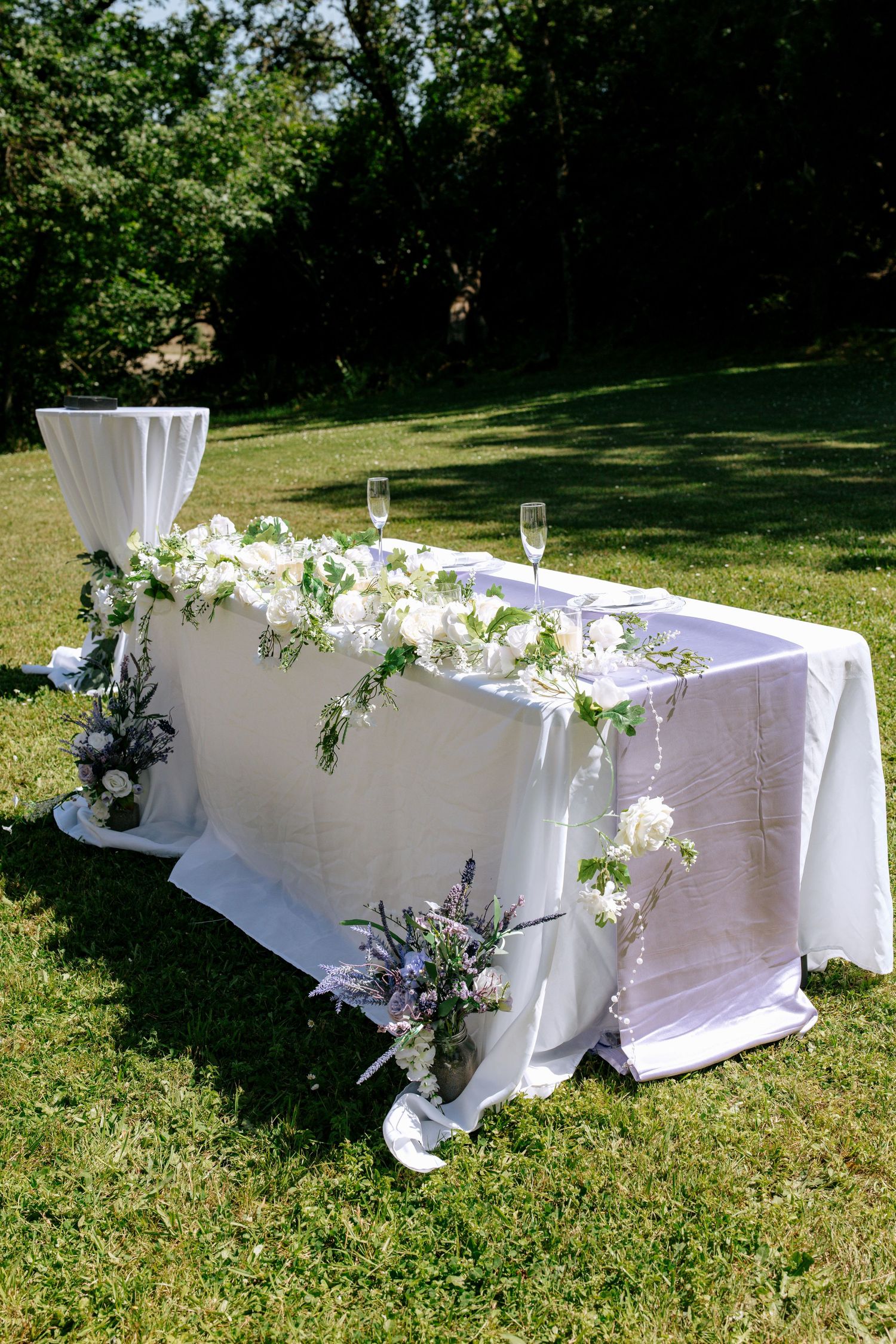 Elegant outdoor wedding sweetheart table draped in white linen adorned with white floral arrangements on green grass.