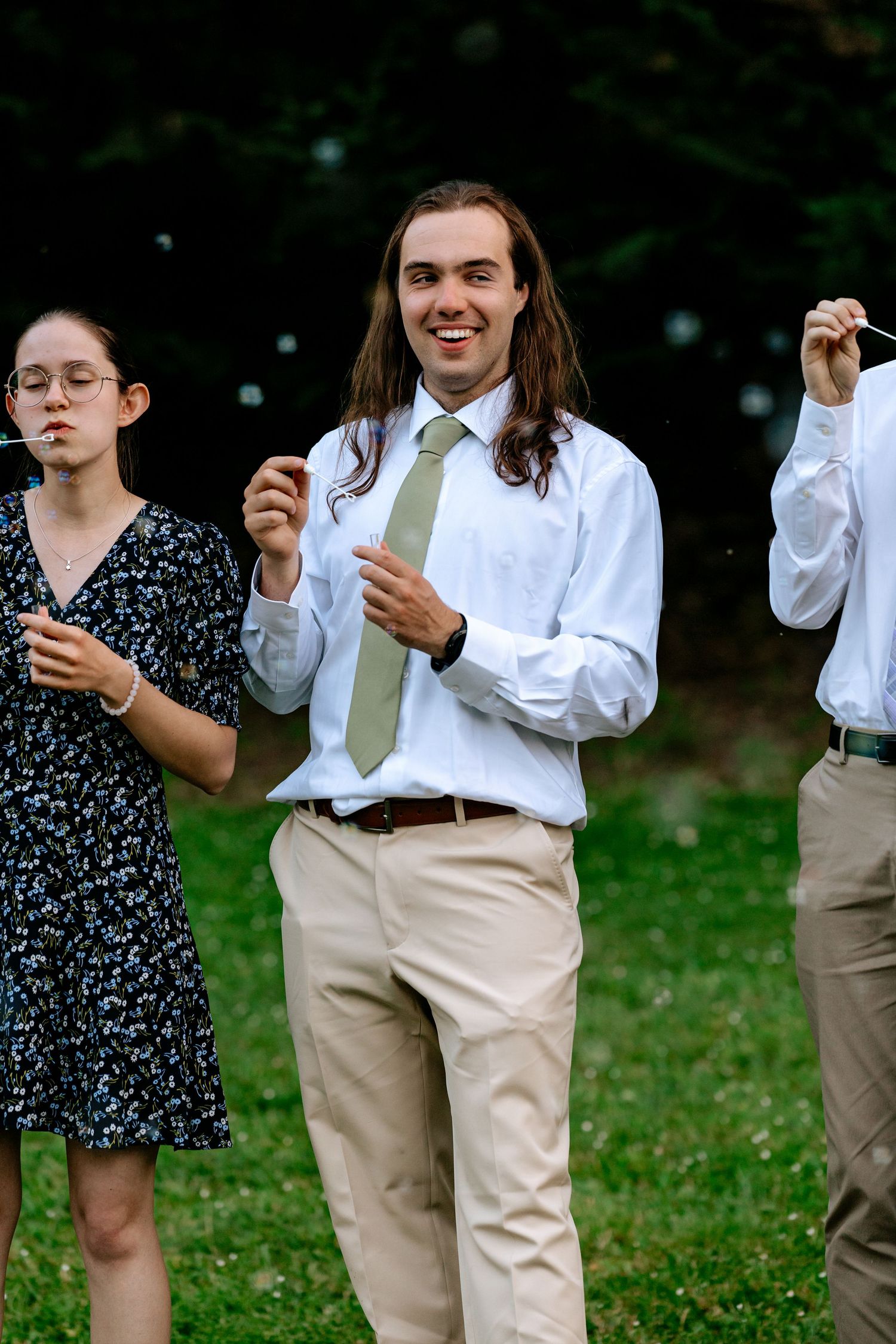 Smiling groomsman in white shirt and sage green tie blows bubbles at outdoor wedding celebration on grass.