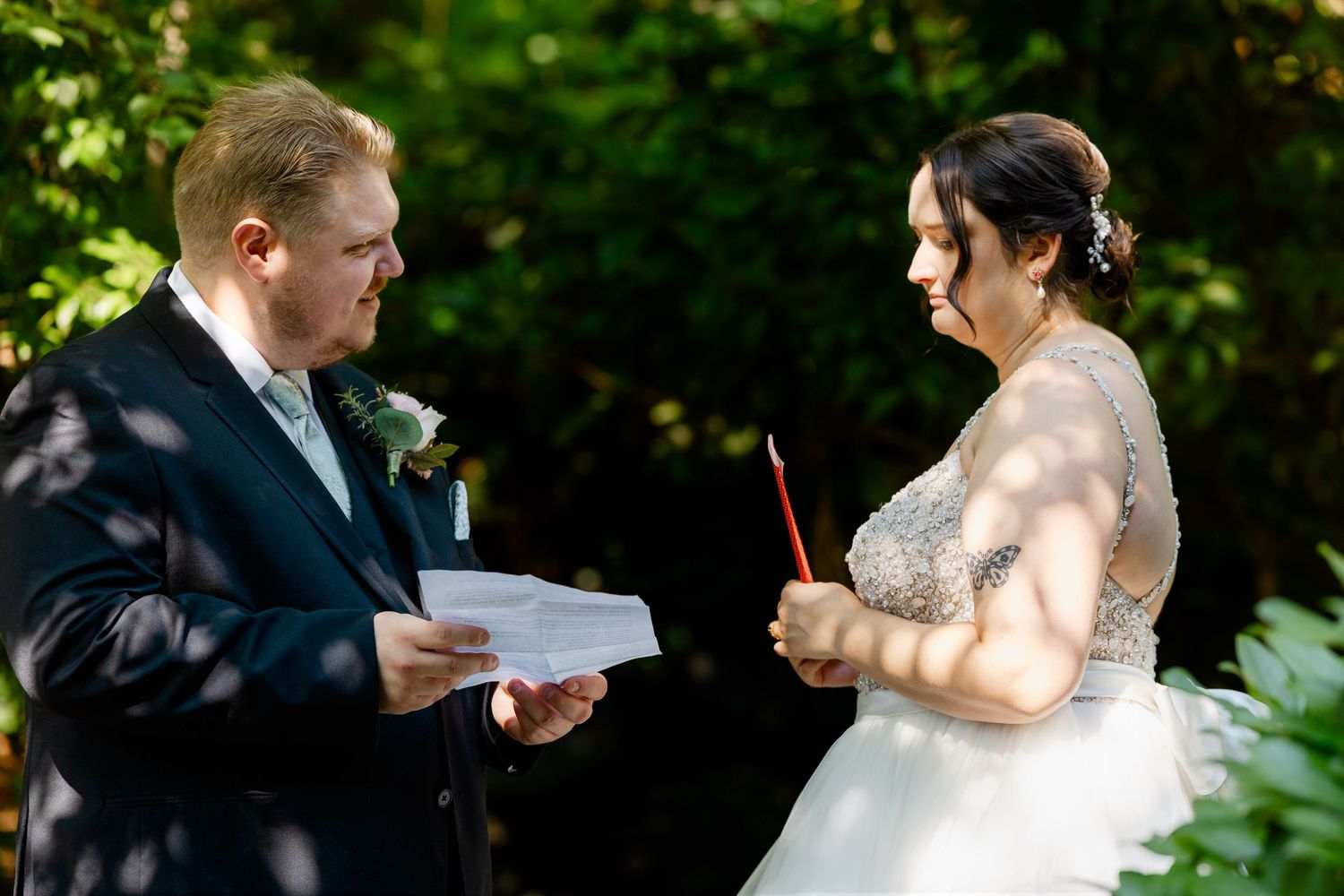 Bride and groom exchange vows outdoors, groom holding papers while bride holds a red pen during wedding ceremony.