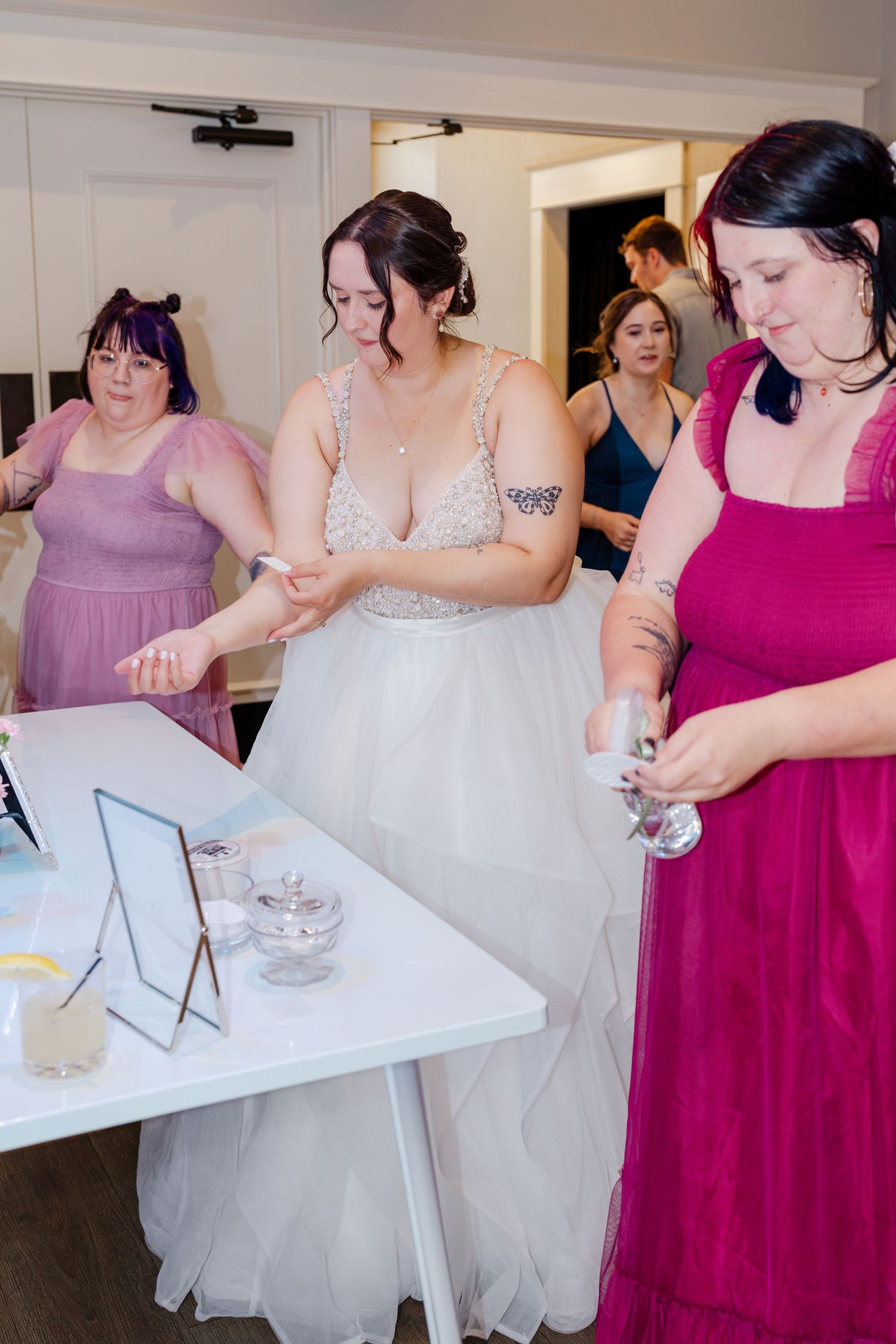 Bride in white gown with bridesmaids in purple and magenta dresses preparing at a white table.