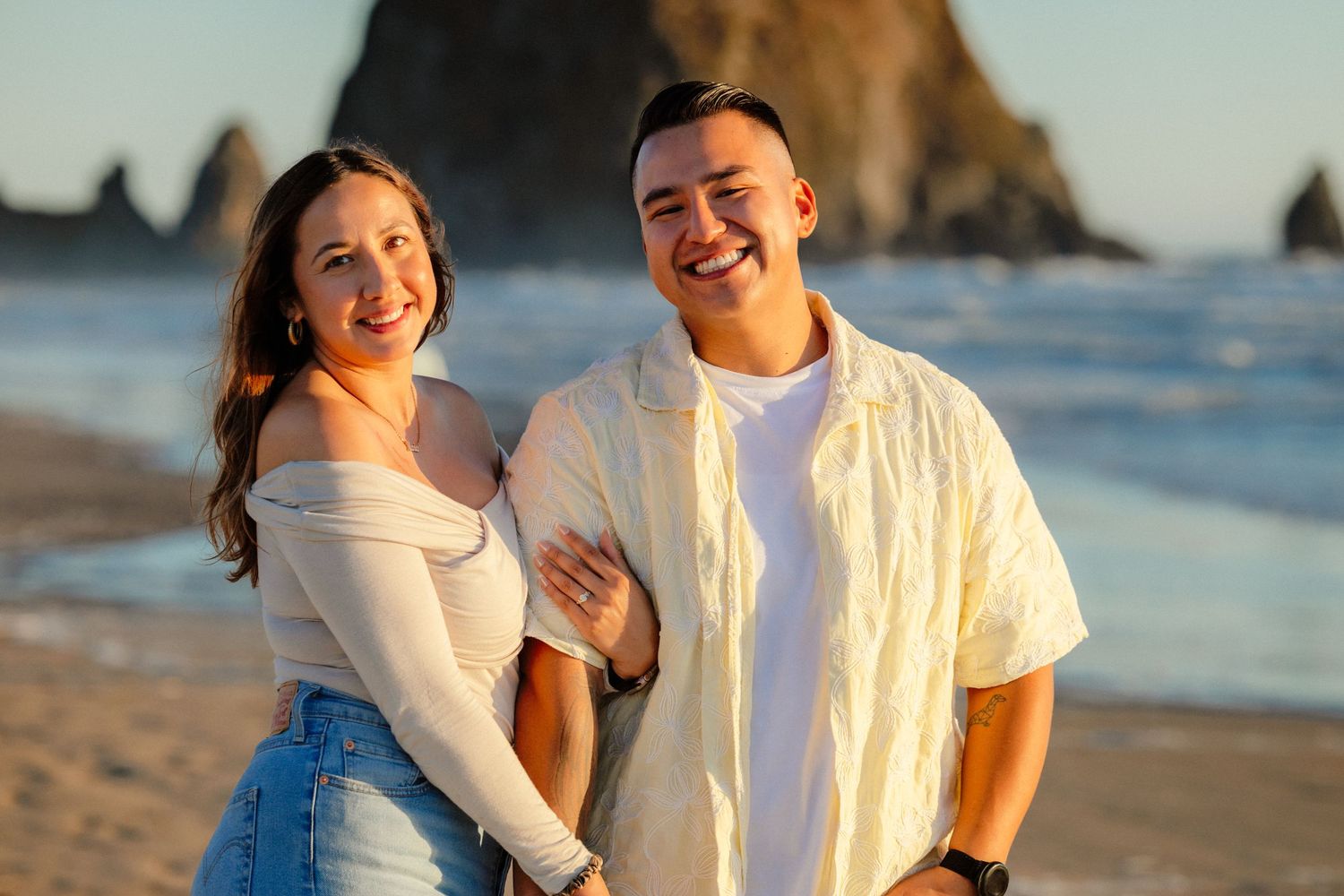 A smiling couple poses together on a sandy beach with ocean waves and a rocky sea stack in the background.