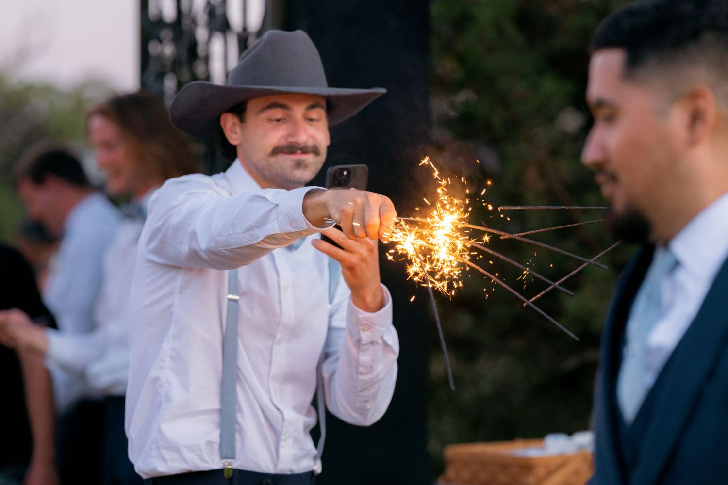 Man in cowboy hat photographs a sparkler at a wedding celebration outdoors.