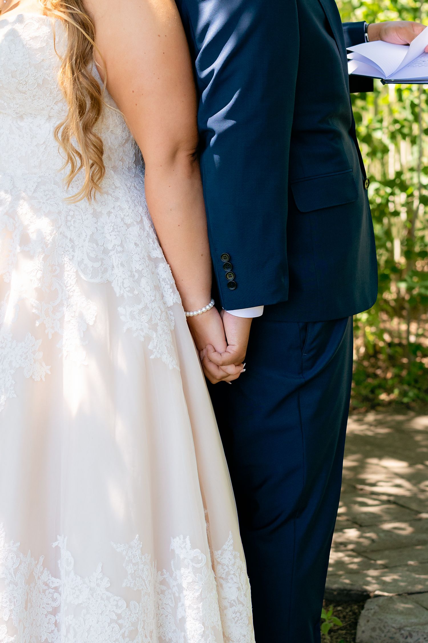 Close-up of bride and groom holding hands during wedding ceremony, bride wearing lace dress with pearl bracelet.