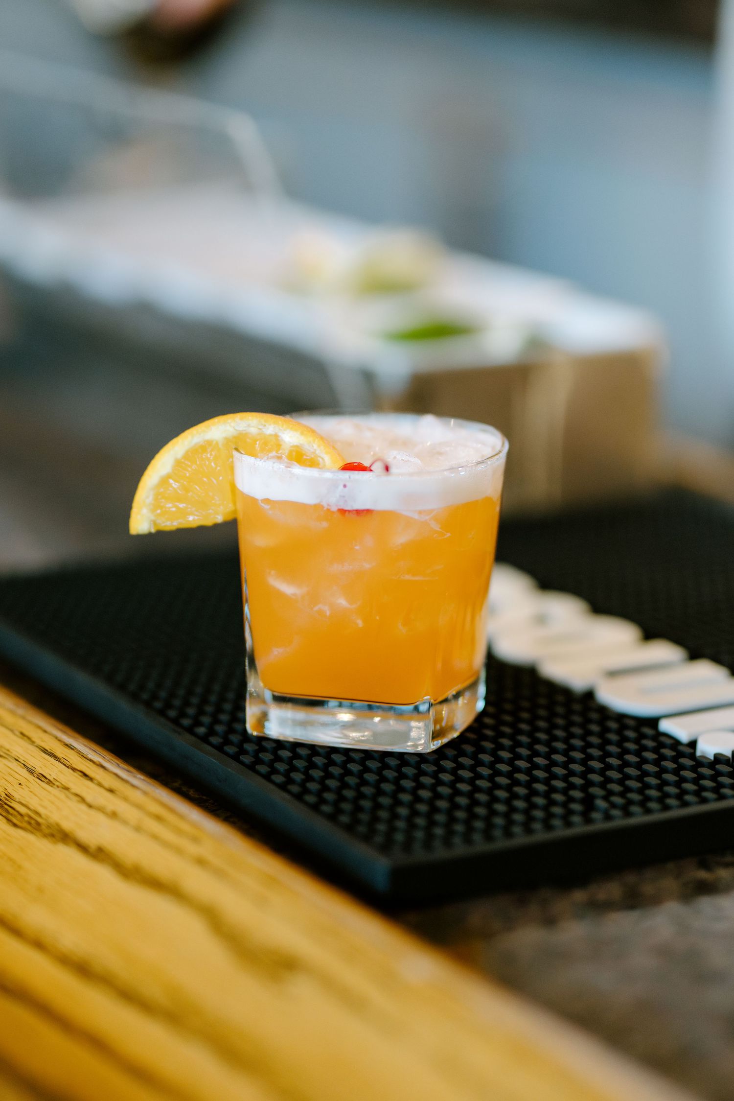 Orange whiskey sour cocktail with lemon slice garnish served on black bar mat at wedding reception.