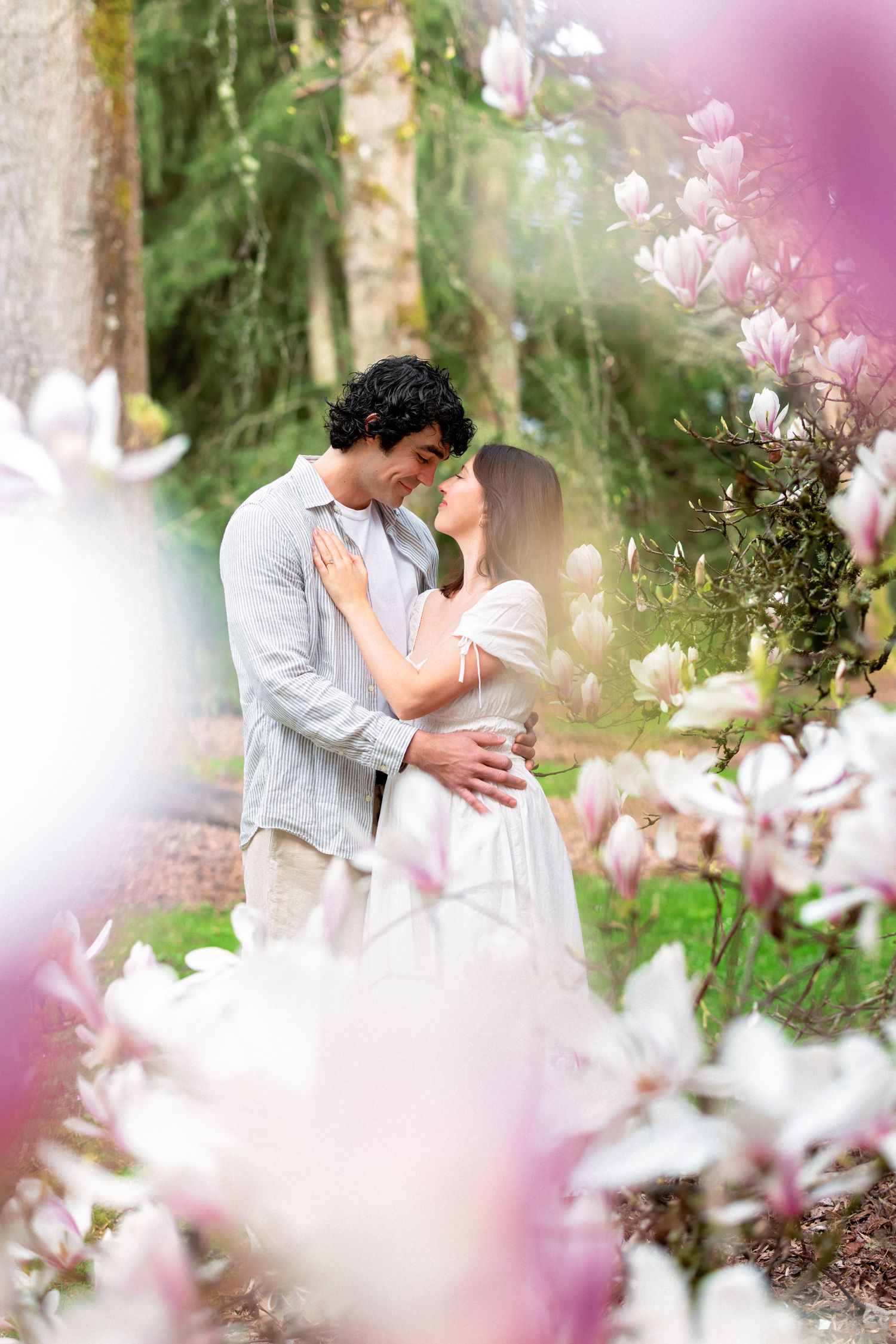 A romantic couple embraces among blooming pink magnolia flowers in a dreamy spring garden setting.