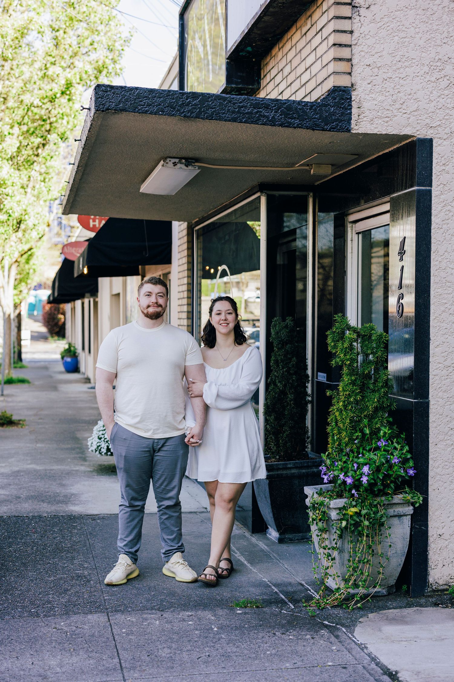 A couple stands together outside a brick storefront with planters and an awning on a sunny day.