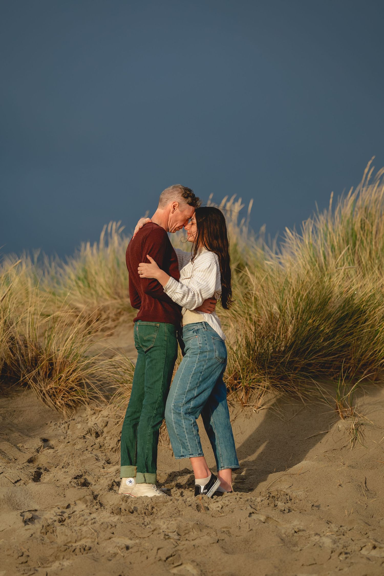 A couple embraces on a sandy dune with tall beach grass and moody blue sky in the background.