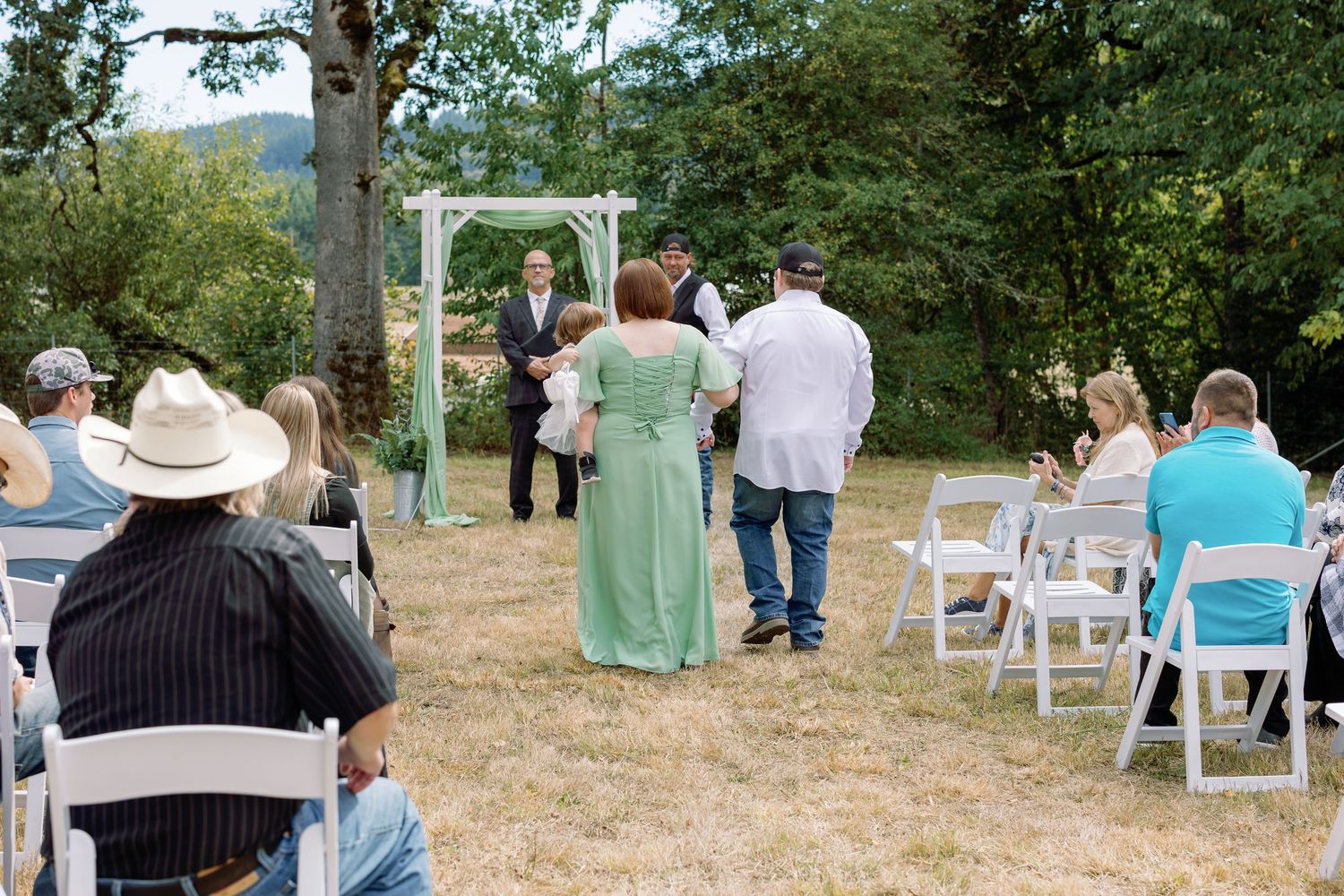 Outdoor wedding ceremony setup with white chairs arranged in rows on a grassy hillside surrounded by trees.