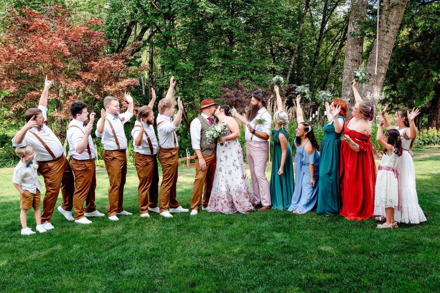 Wedding party poses playfully in a line on green grass during outdoor summer celebration.