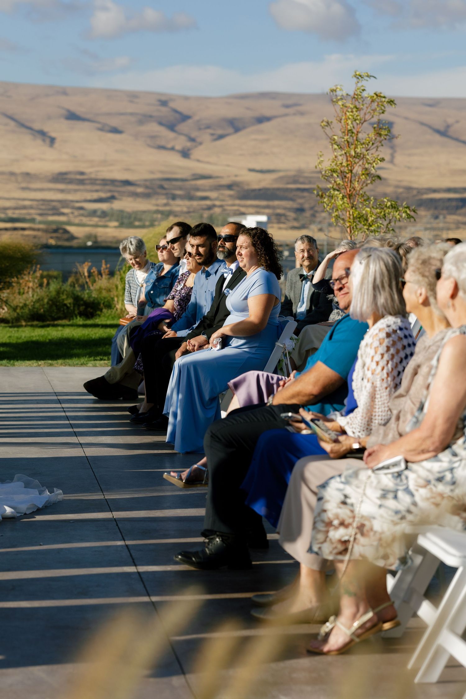 Wedding ceremony guests seated in white chairs overlooking a scenic mountain landscape on a sunny day.