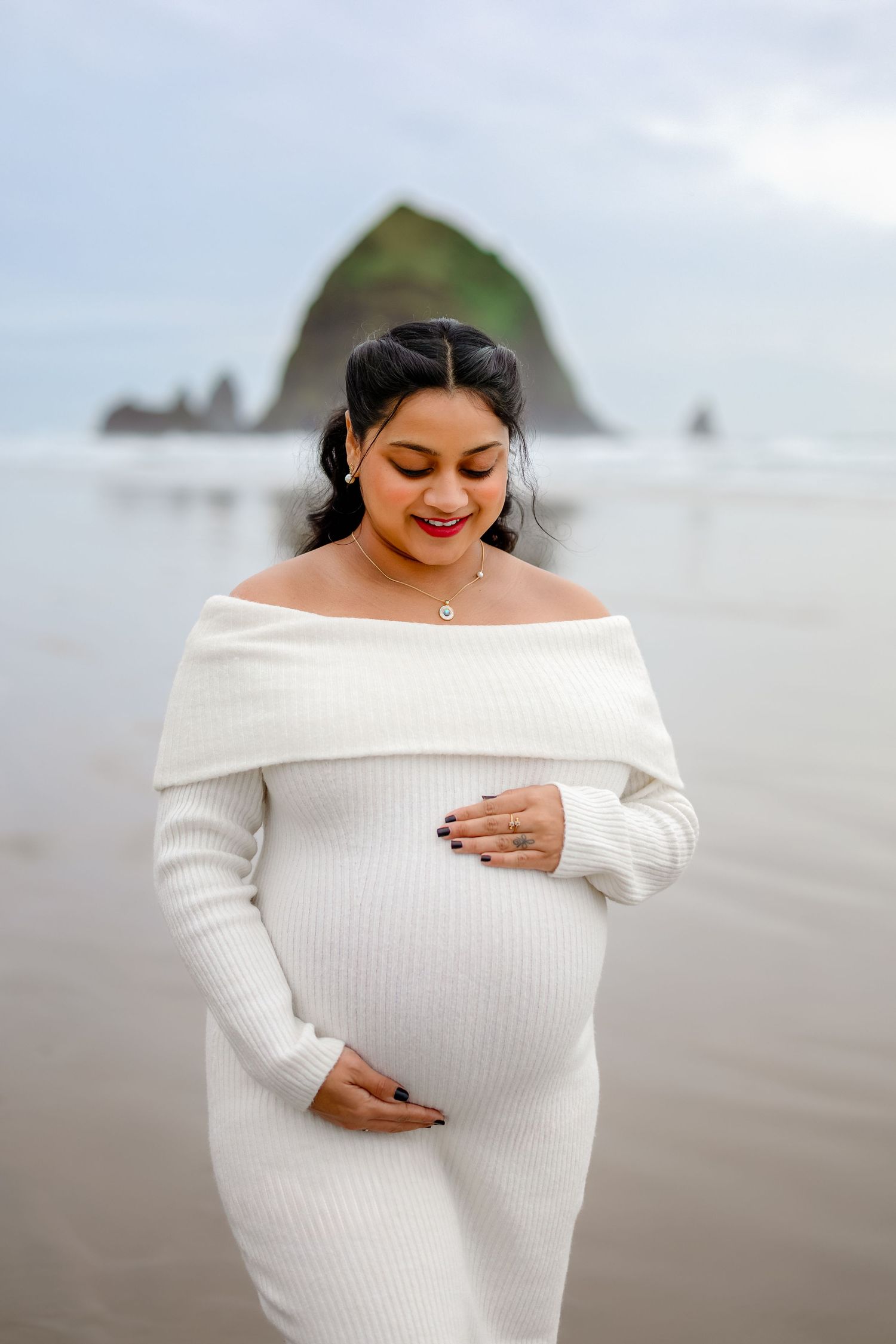 A person wearing a white off-shoulder sweater smiles while looking down at their phone on a cloudy beach.