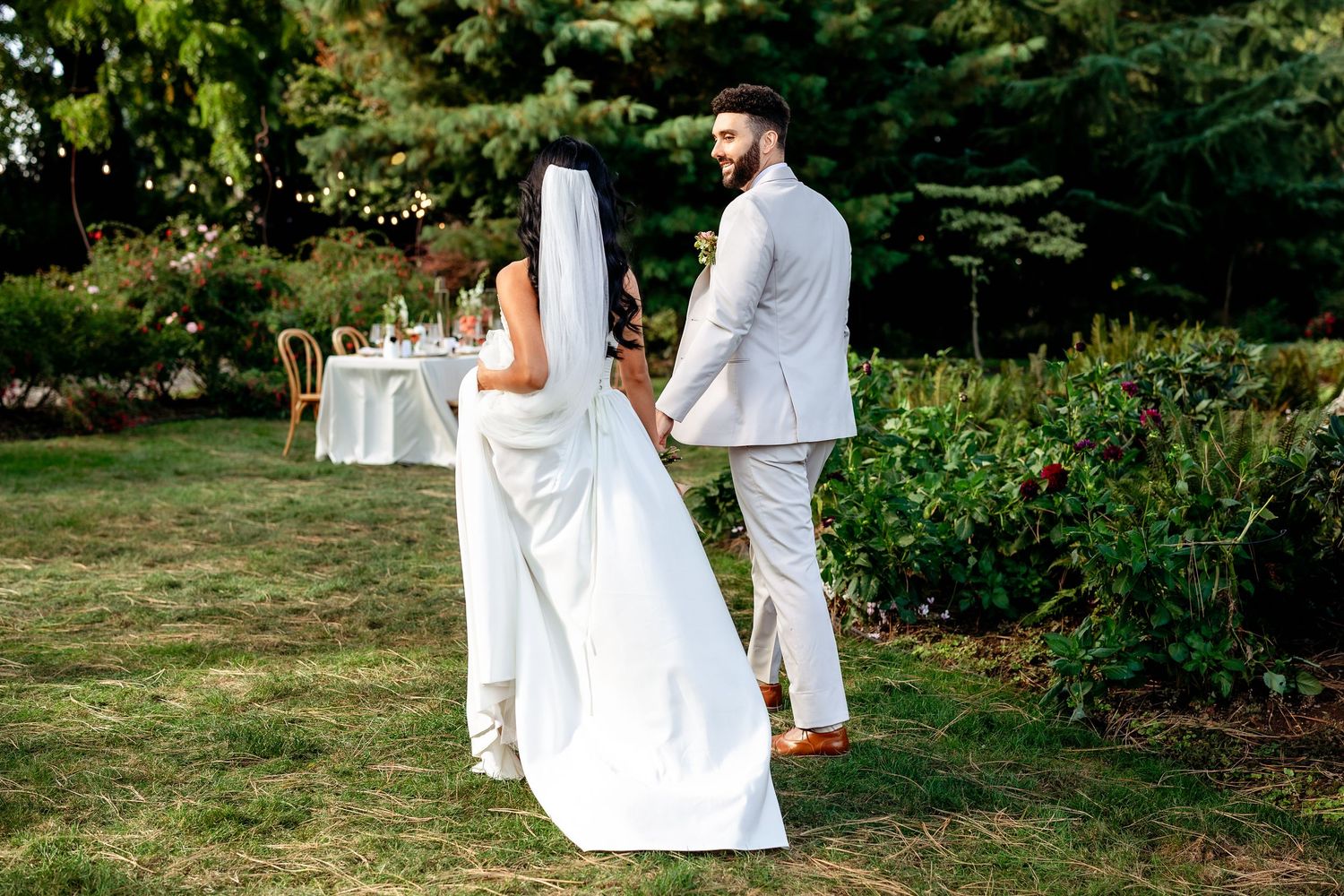 Wedding couple poses in elegant white outfits against lush garden backdrop on sunny day.