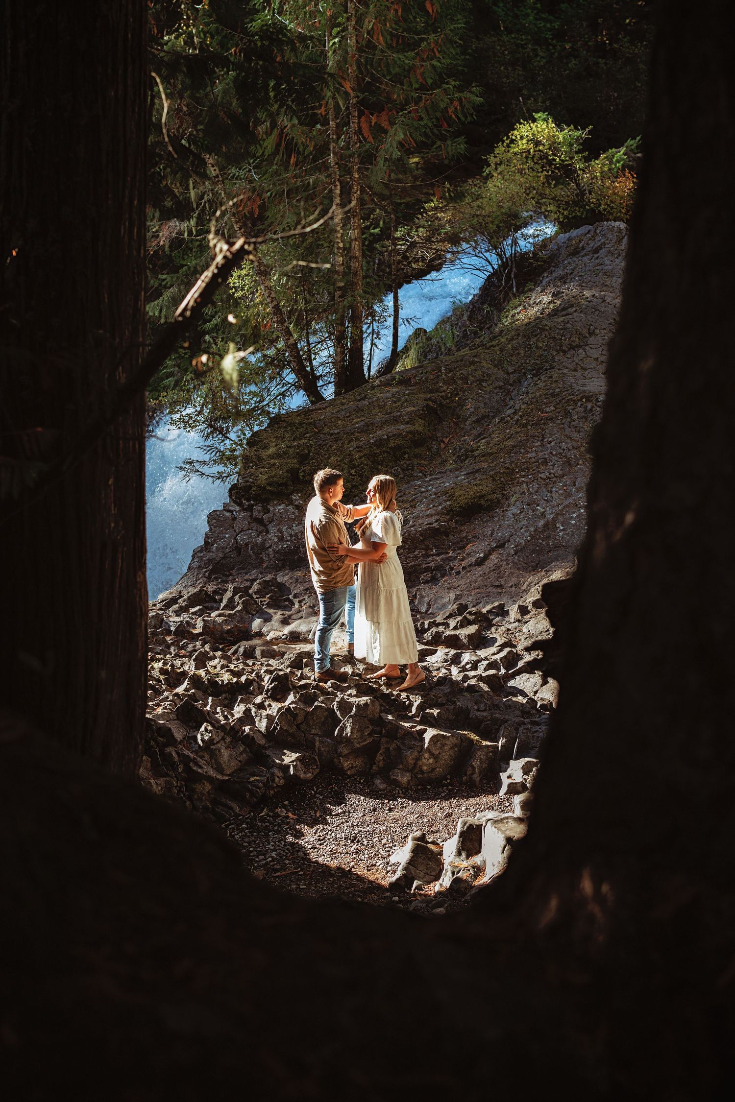 A couple walking through a rocky forest path with sunlight filtering through the trees.