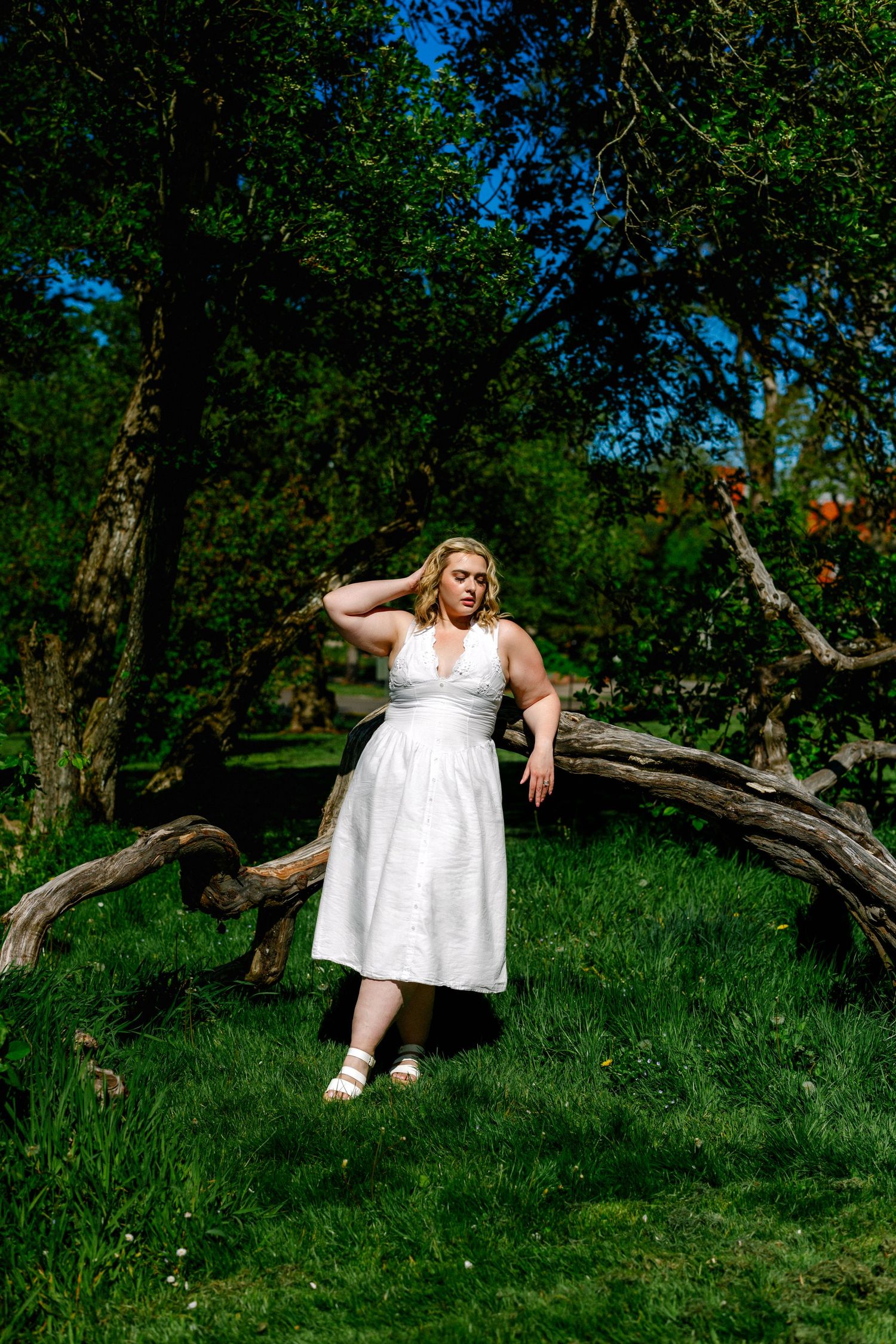 Plus-size woman in white halter midi dress posing against fallen tree trunk in lush green park.