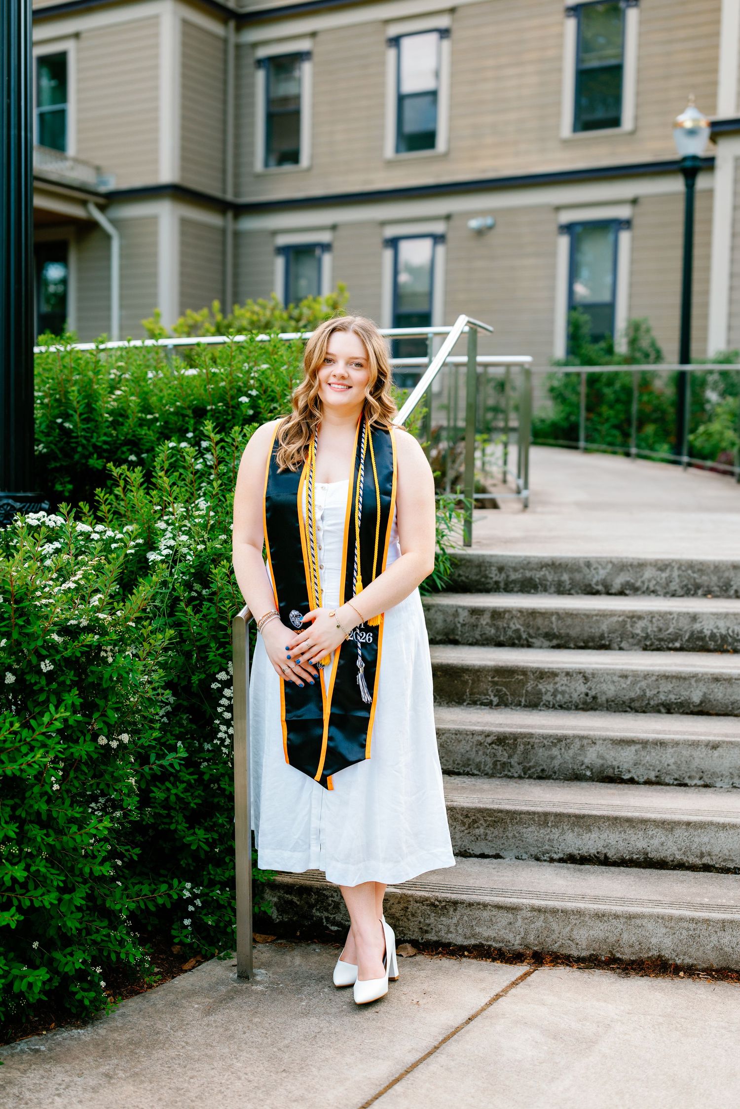 Female graduate in white dress with black and gold stole standing near steps and greenery.