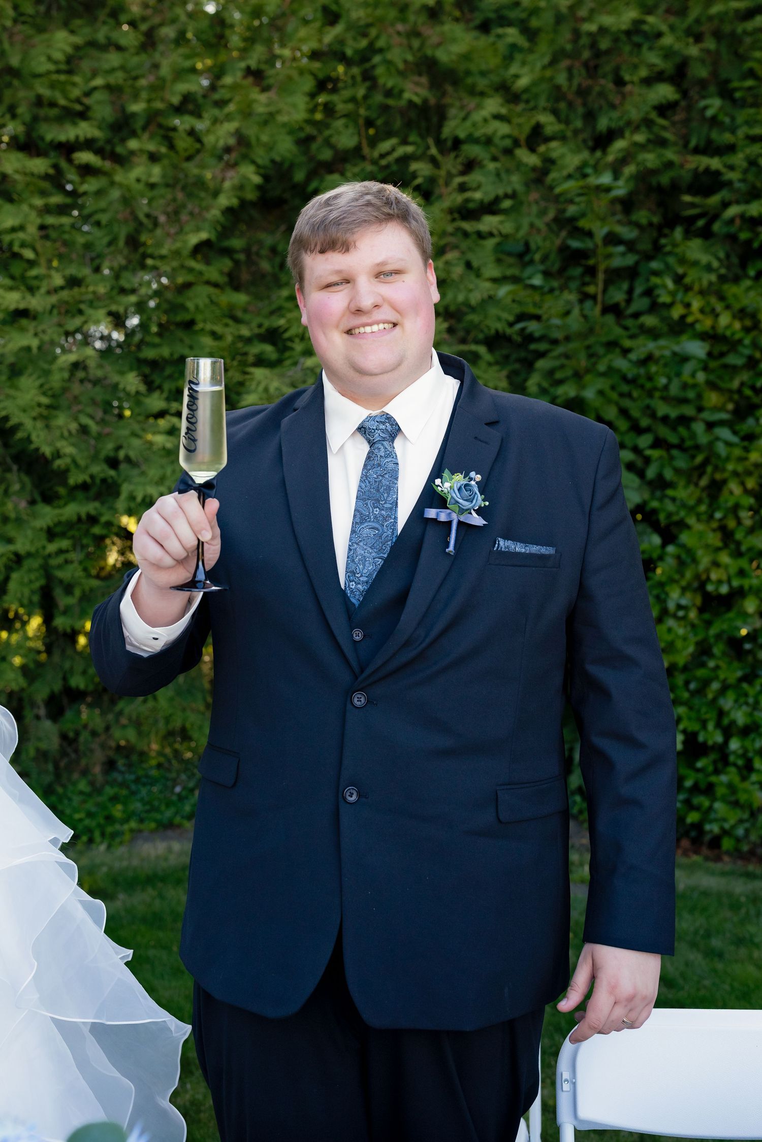Smiling groom in navy suit raising champagne glass with boutonniere at outdoor wedding reception.