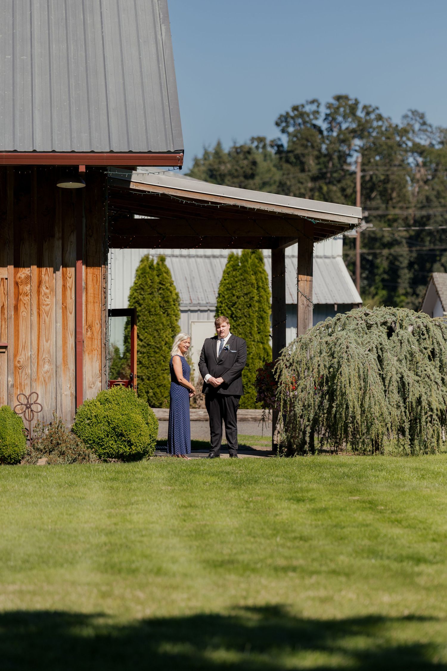 A couple stands together on a green lawn beside a rustic wooden barn venue on a sunny day.