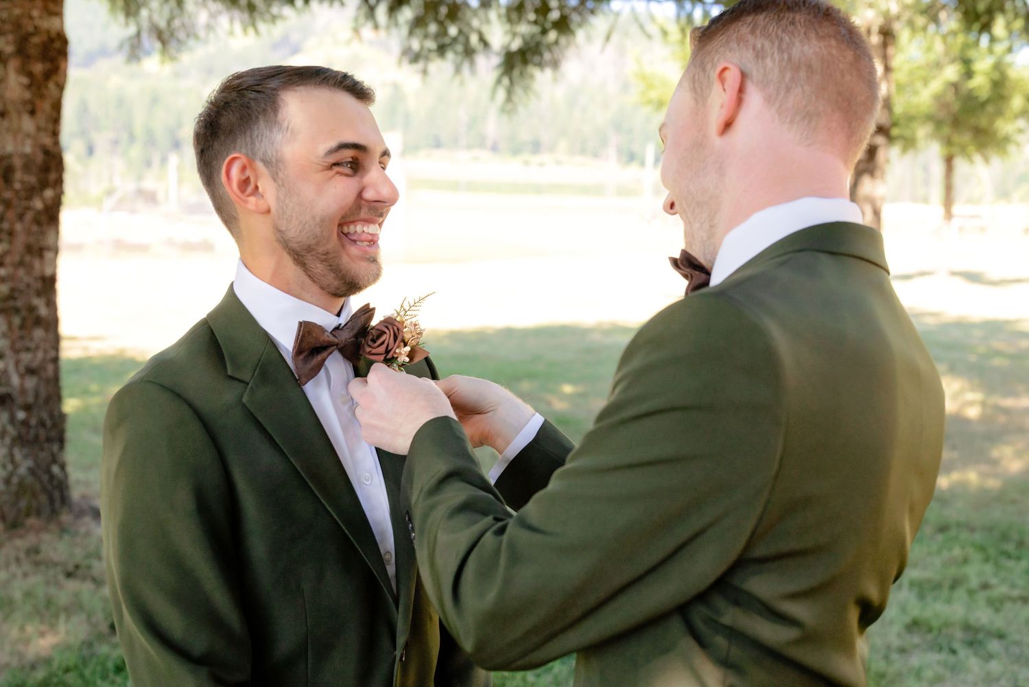 Groomsman adjusting boutonniere on smiling groom in olive green suit outdoors at wedding.