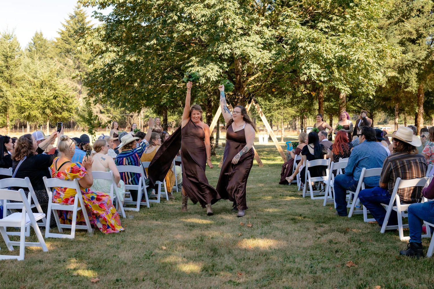 Two bridesmaids in brown dresses celebrating in the aisle at an outdoor wedding ceremony.