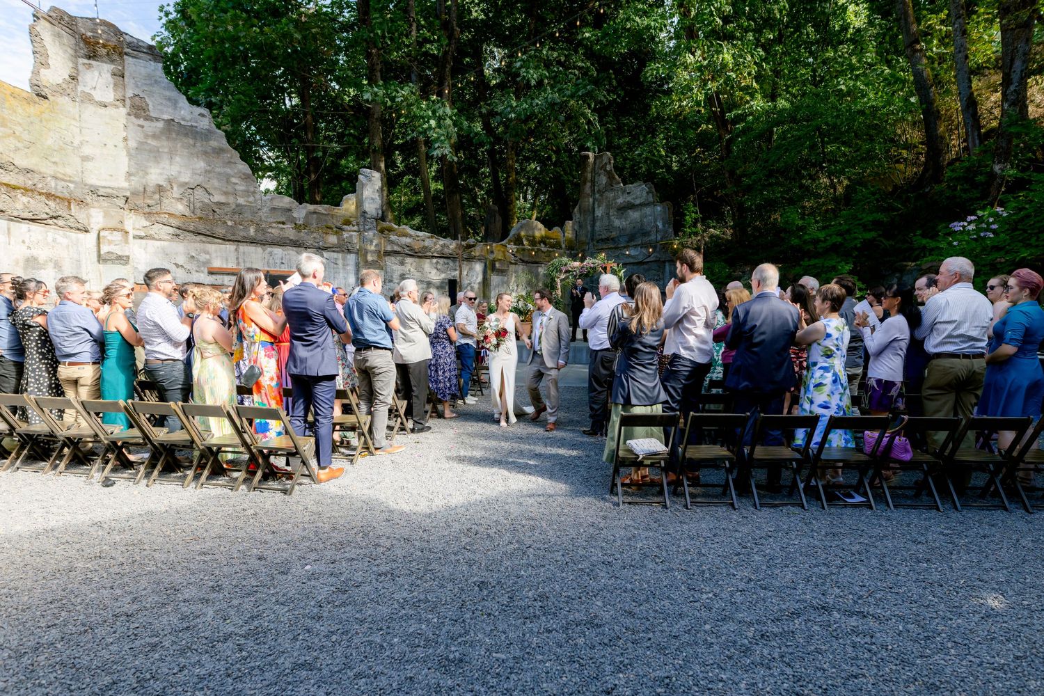 Wedding ceremony guests seated in wooden chairs at outdoor stone ruins venue surrounded by tall green trees.