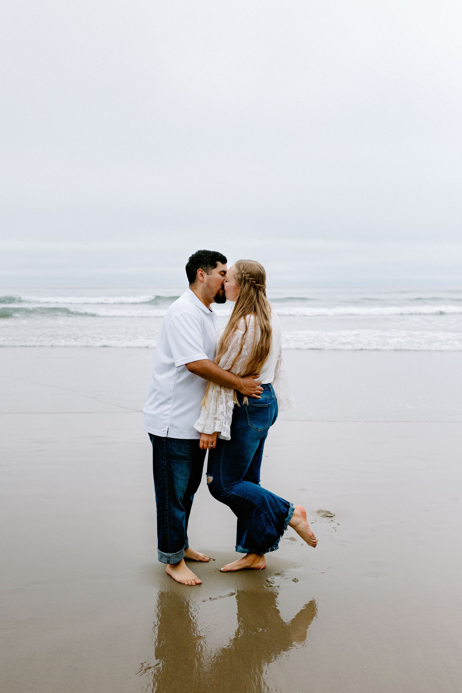 Couple sharing a kiss on a wet sandy beach with ocean waves and cloudy sky behind them.