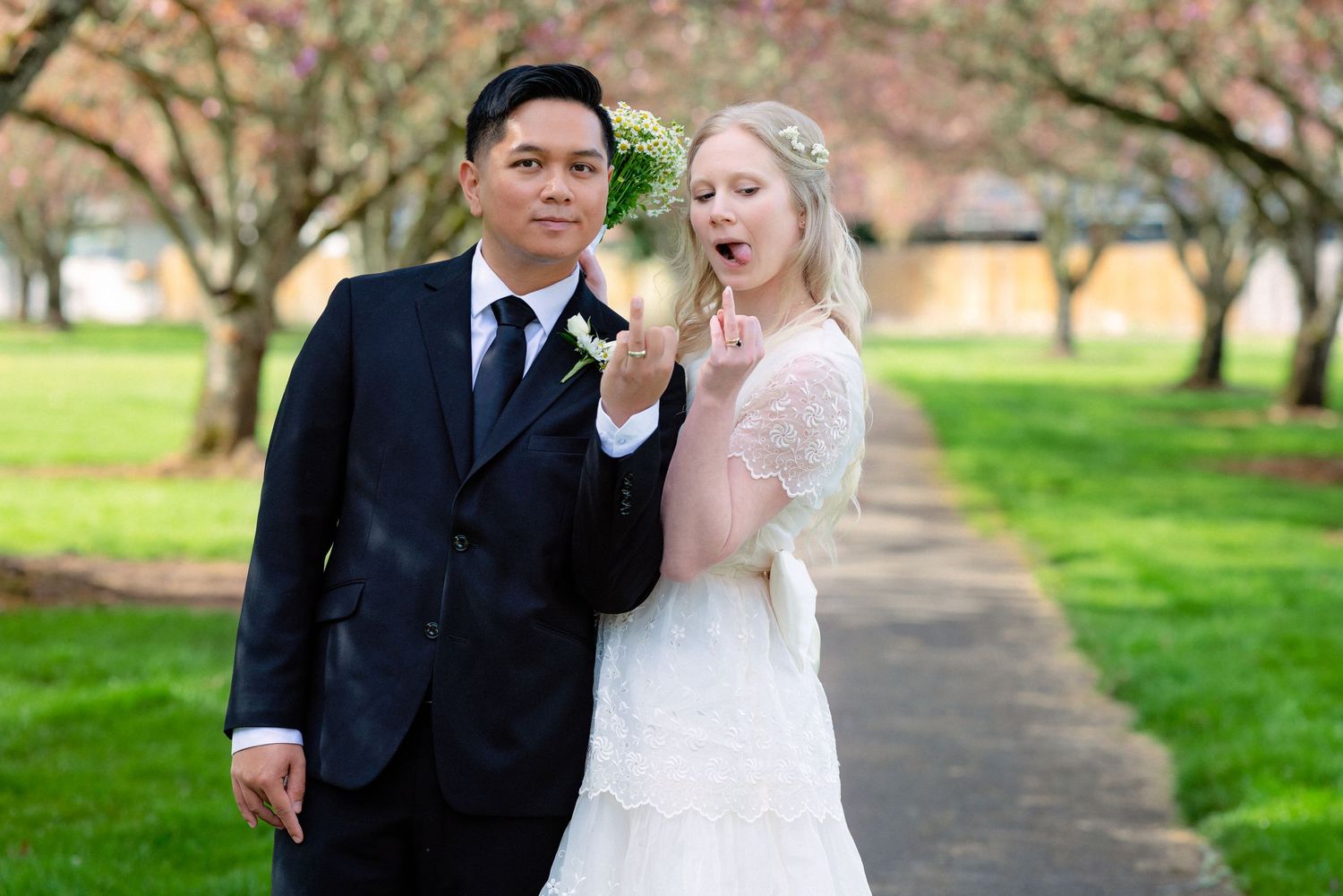 A groom in a navy suit and bride in a white dress pose playfully together outdoors among spring blossoms.
