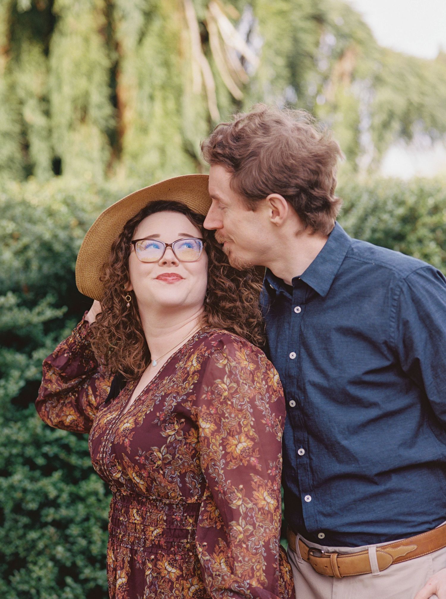 A couple in coordinated outfits poses affectionately against a green garden backdrop.