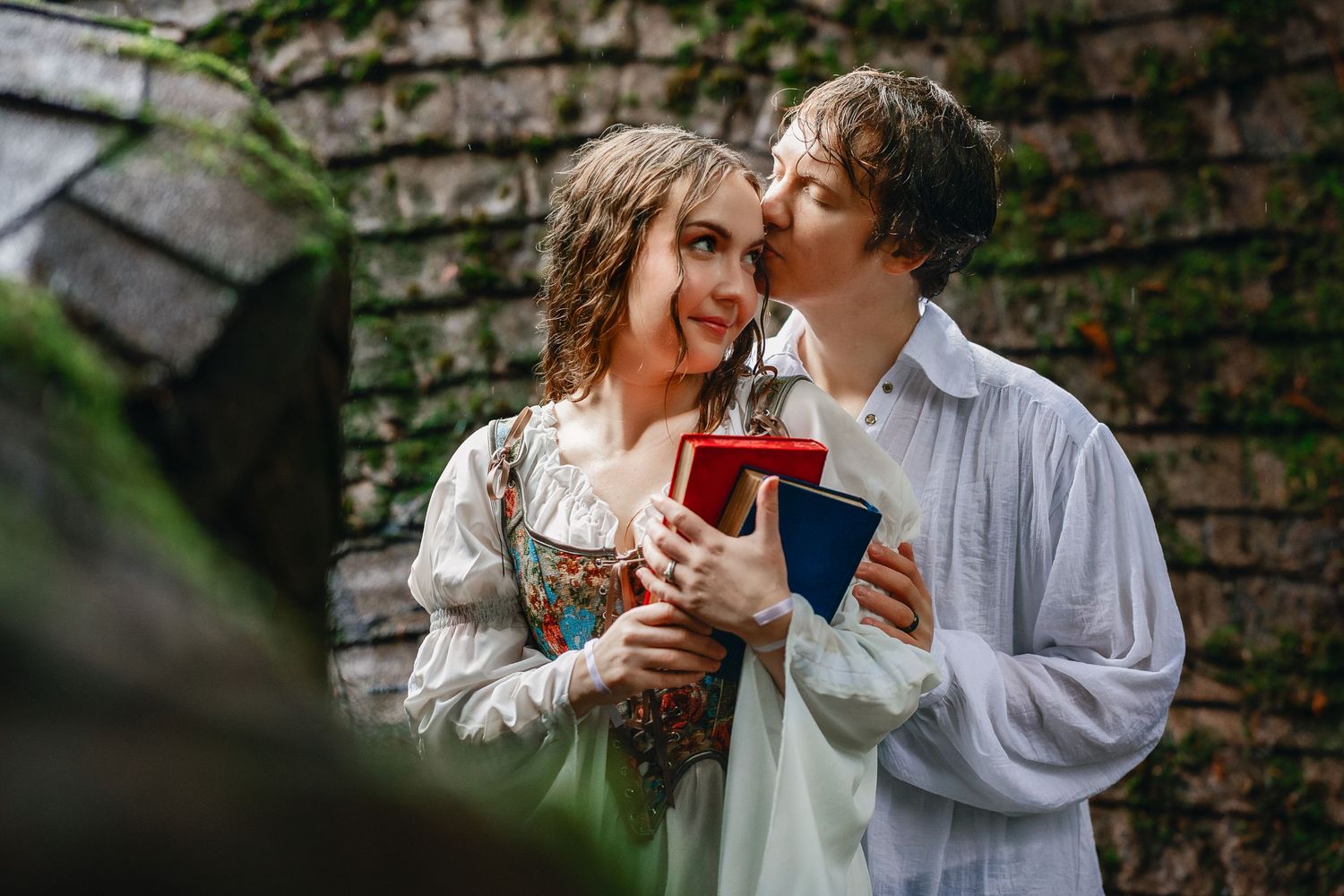 A couple shares a romantic moment while holding drinks in front of a rustic brick wall with greenery.