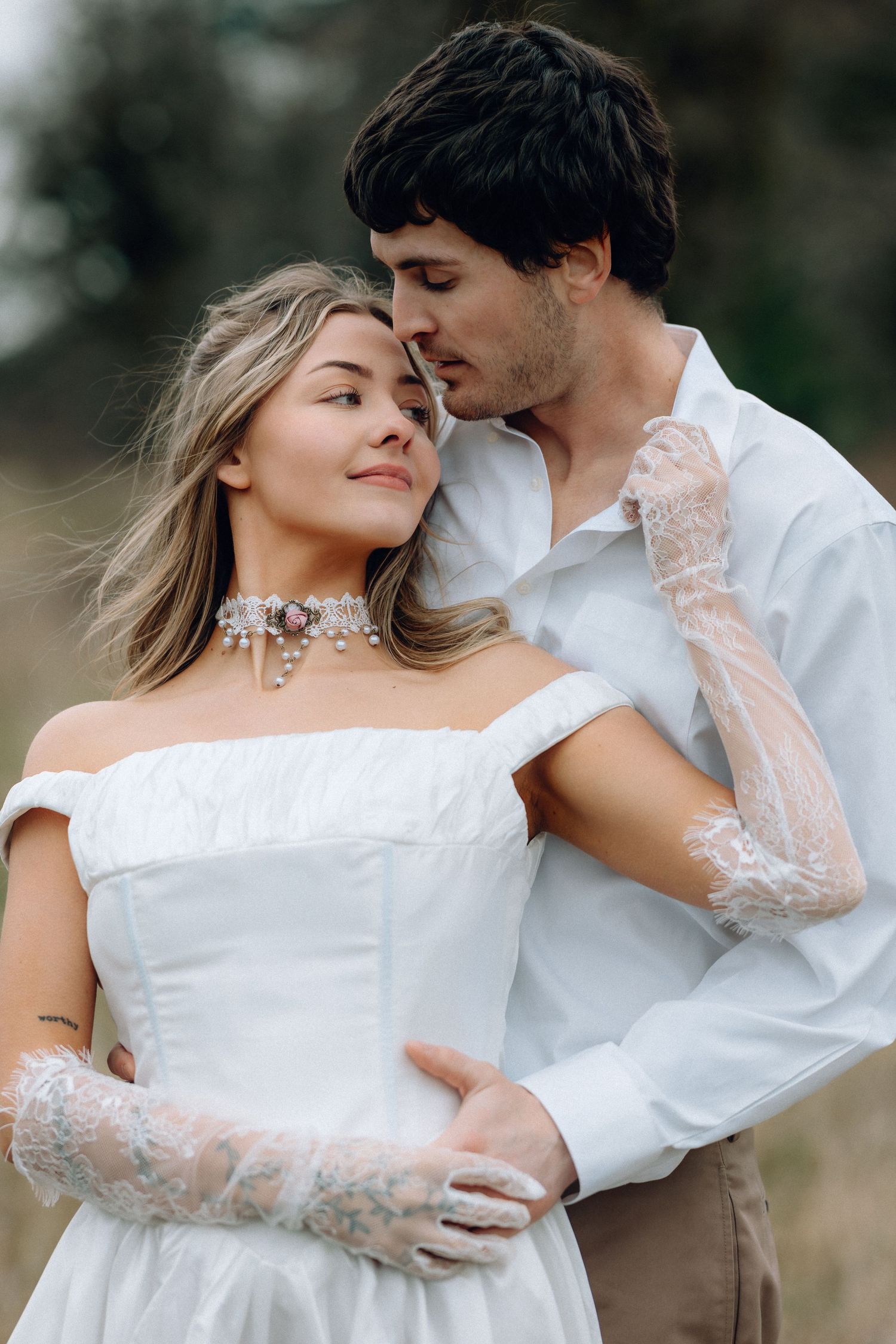 Romantic wedding couple embracing in white attire against a natural outdoor backdrop.