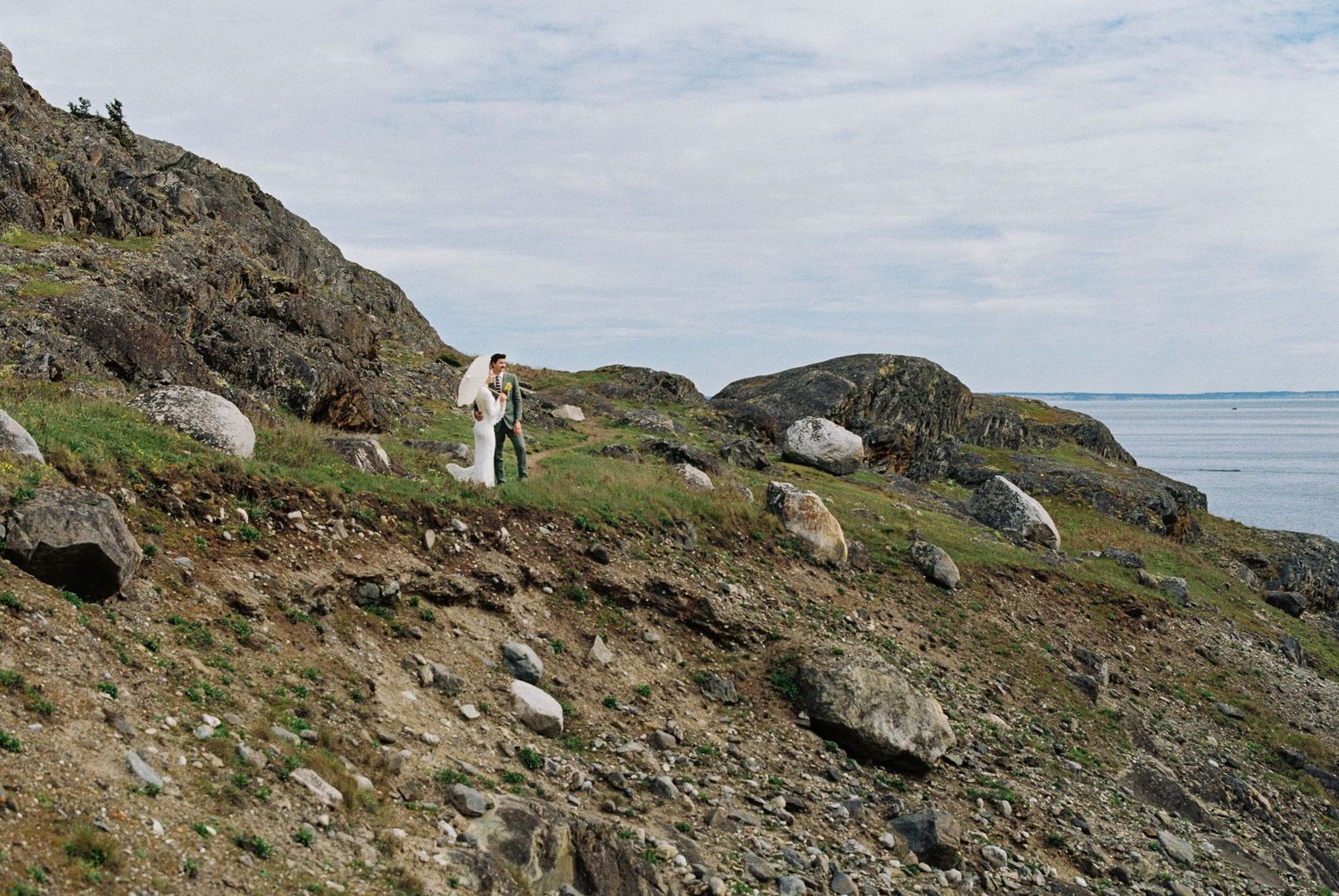 Person hiking on a rocky coastal hillside with boulders and calm ocean waters visible in the background.