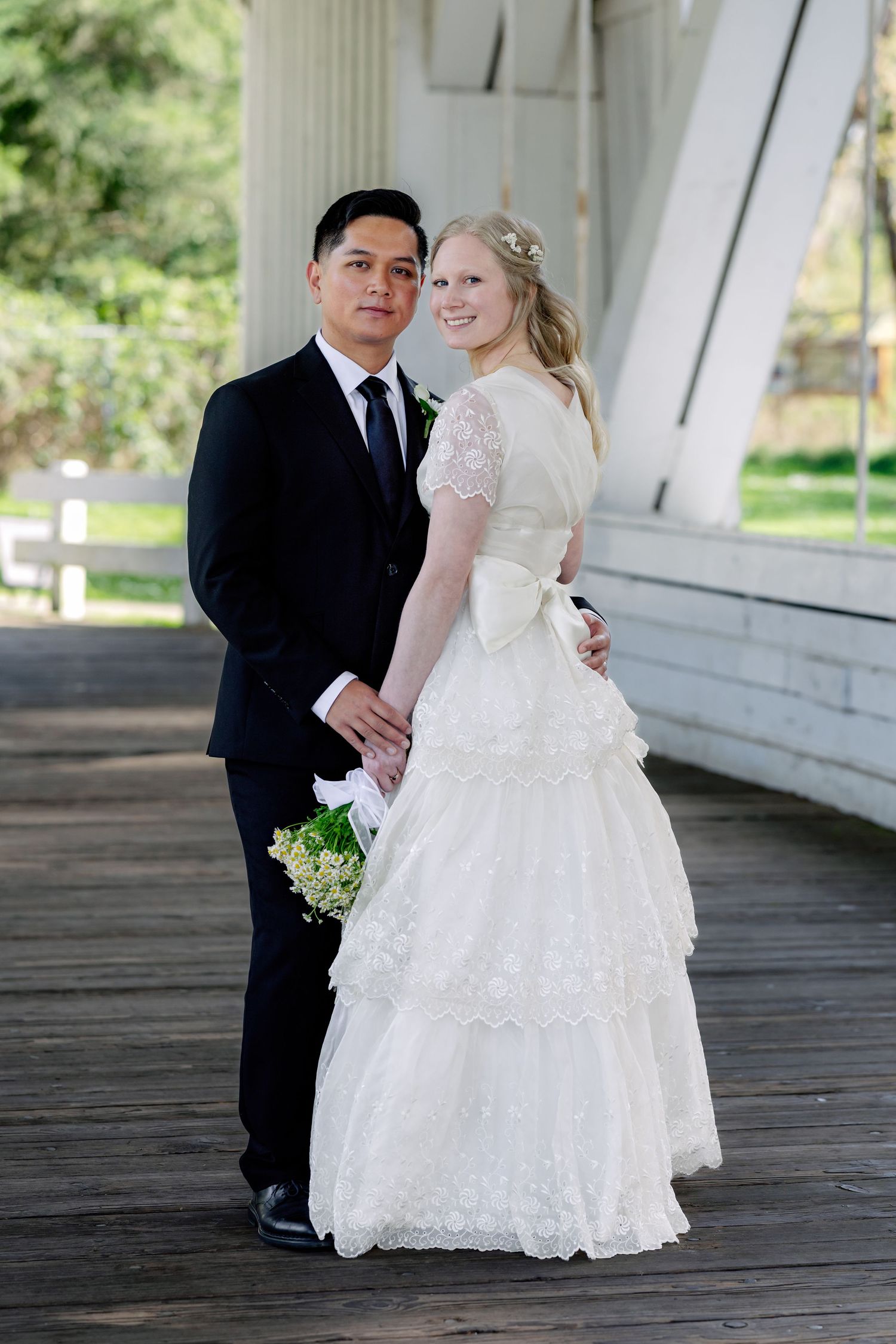 Newlywed couple poses together on wooden covered bridge, bride in white lace gown holding bouquet.