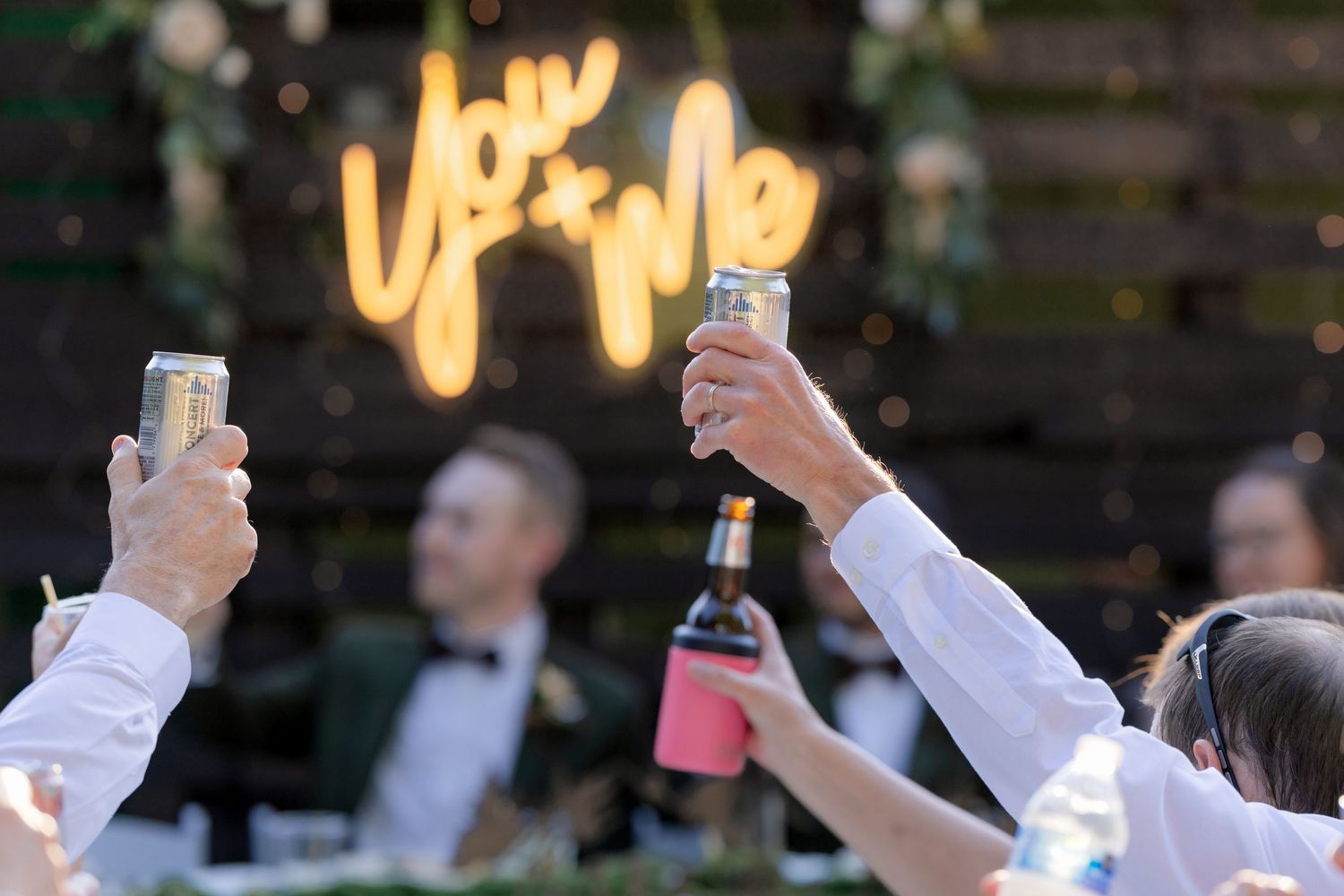Wedding guests raise cans and bottles in a toast beneath a glowing You and Me neon sign at reception.
