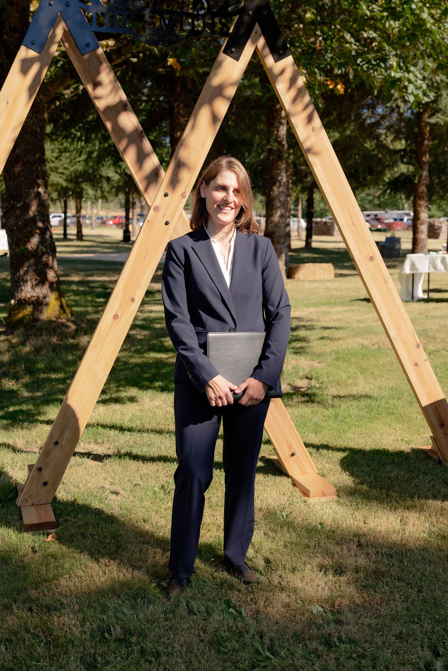 Female officiant in navy suit holding a book stands under a wooden triangular arch in a park.
