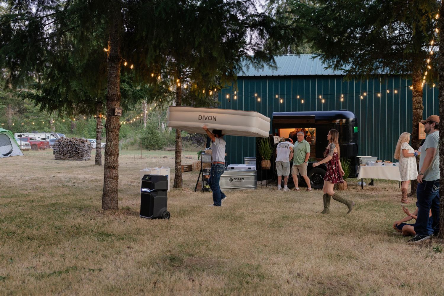 Wedding guests gather near a vintage trailer bar with string lights at a rustic outdoor evening reception.