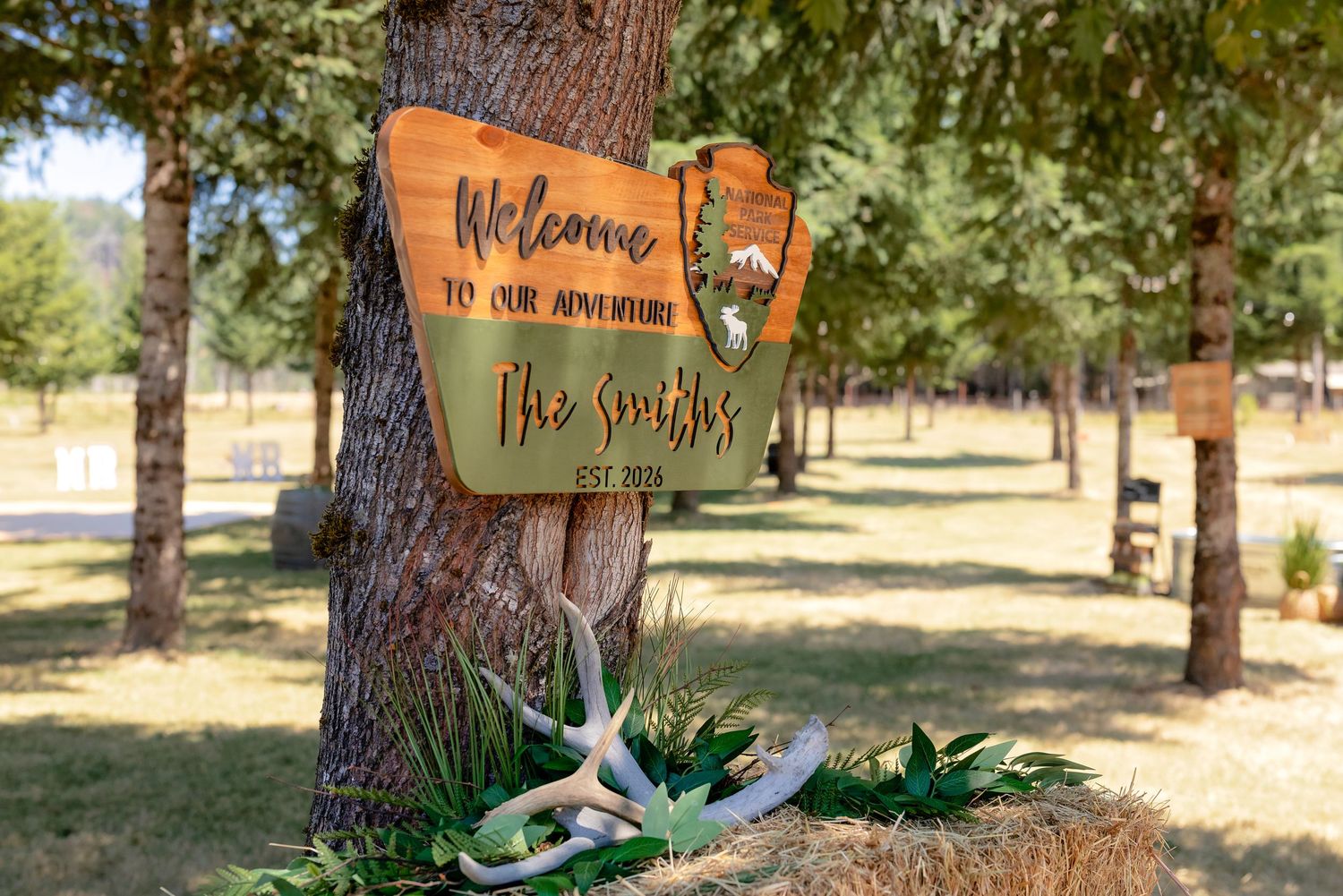 Rustic wooden welcome sign for The Smiths EST. 2024 on a tree, decorated with antlers and hay bale outdoors.