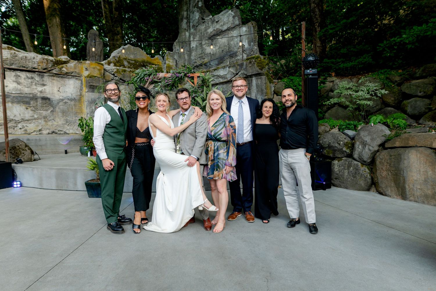 Wedding party group photo in front of mossy rock wall outdoor venue.