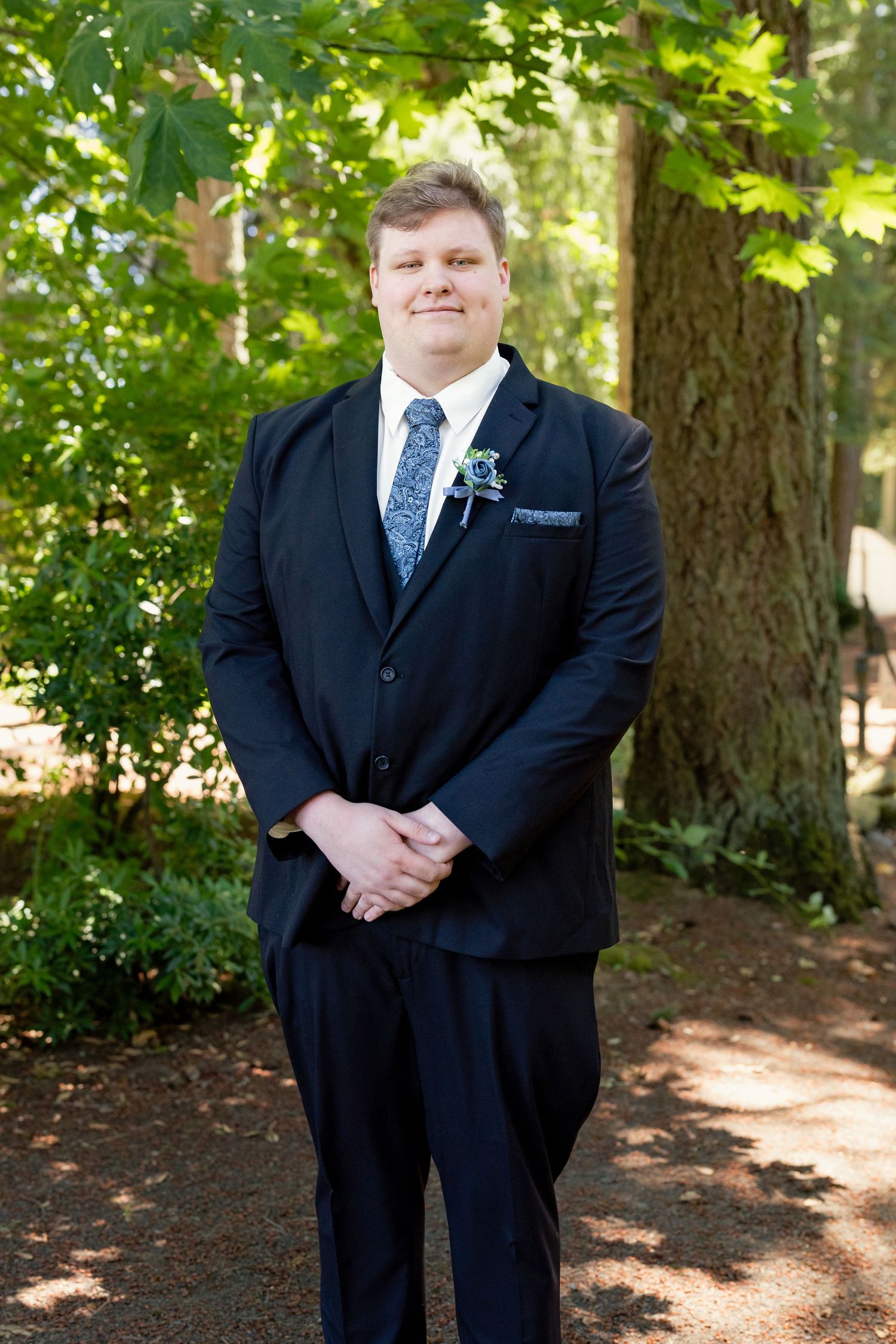 Groom in a navy suit with blue floral tie and boutonniere posing in a lush green forest setting.