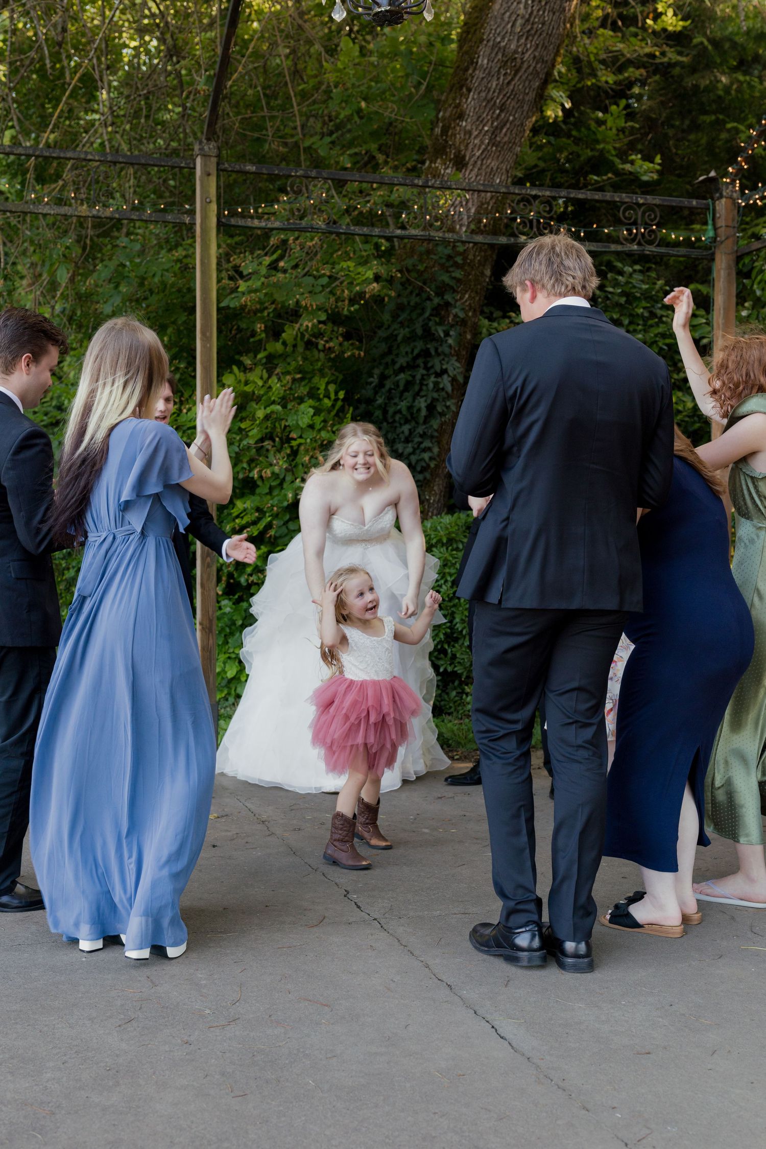 Bride dancing with flower girl and wedding guests on outdoor patio surrounded by greenery.