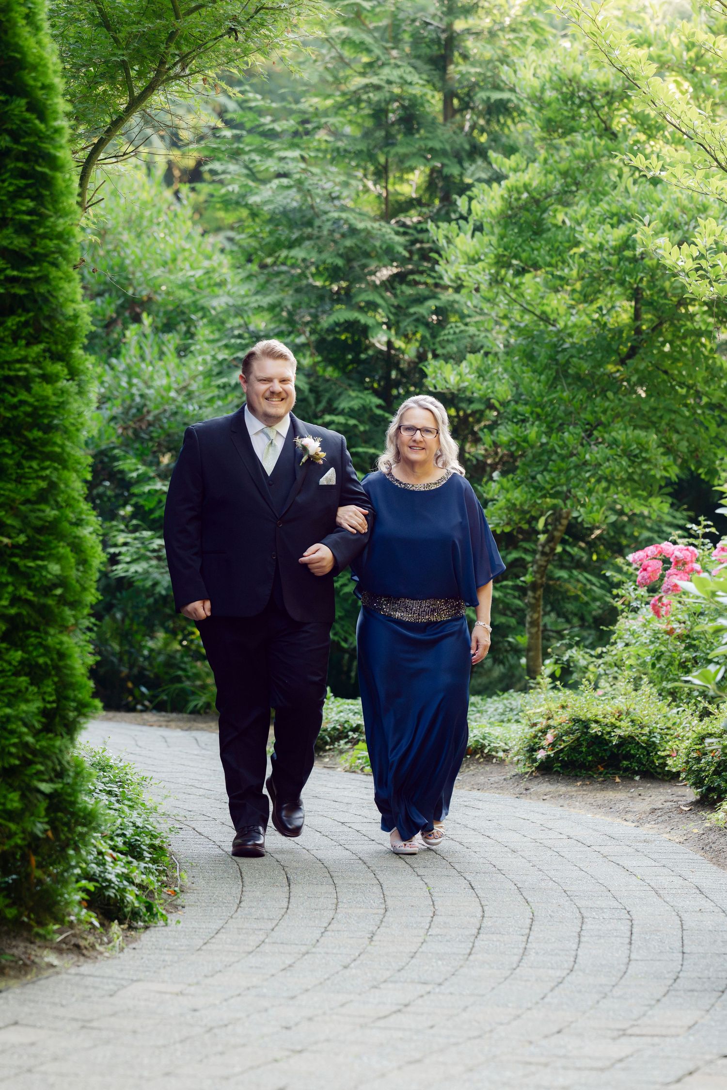 Groom in black tuxedo walking with his mother in a navy dress along a garden pathway.