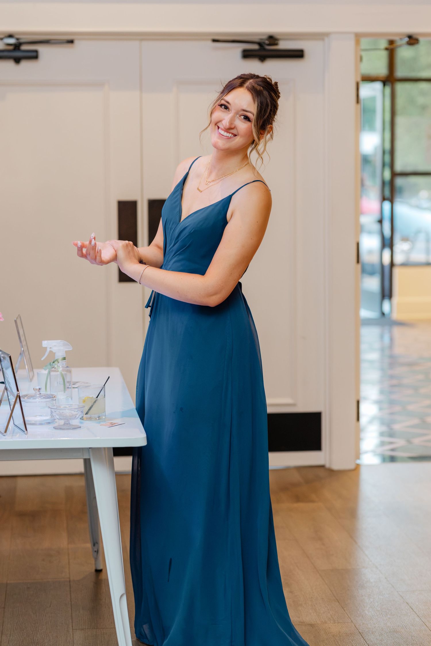 Smiling bridesmaid in teal blue gown standing near a table in a bright elegant indoor venue.
