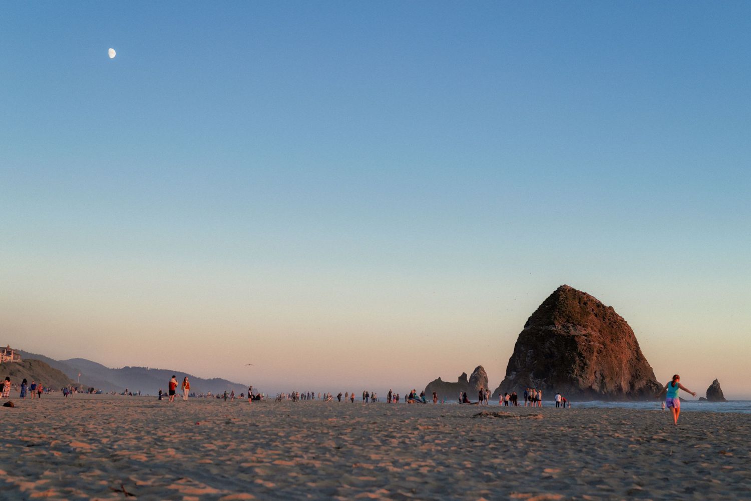Sunset at Cannon Beach with Haystack Rock and people strolling on the sandy shore under a clear sky with the moon visible.